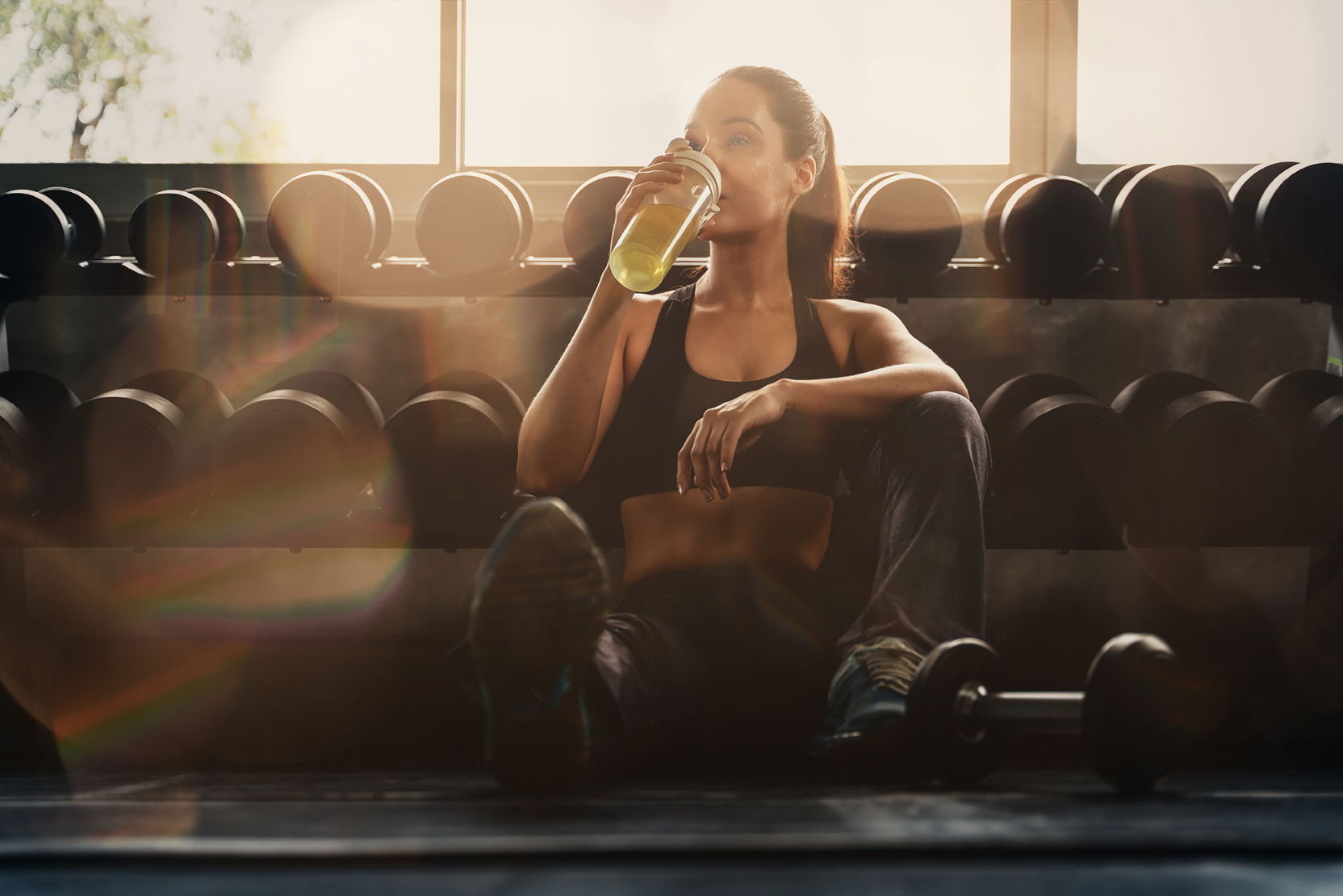Woman sitting in a gym, drinking from a bottle, surrounded by weights.