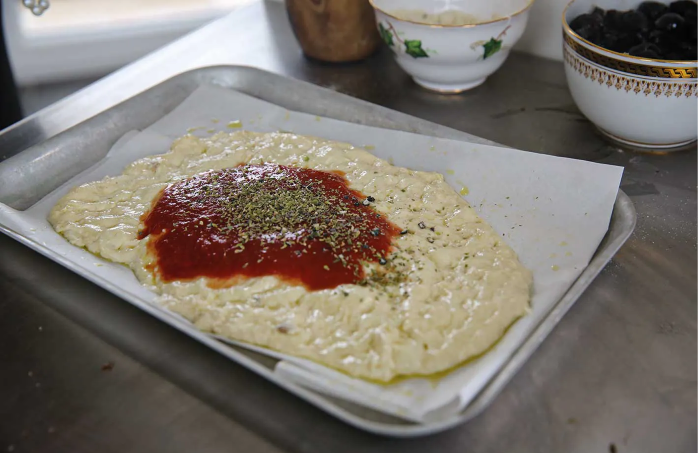 Dough spread on a tray with tomato sauce and herbs on top.