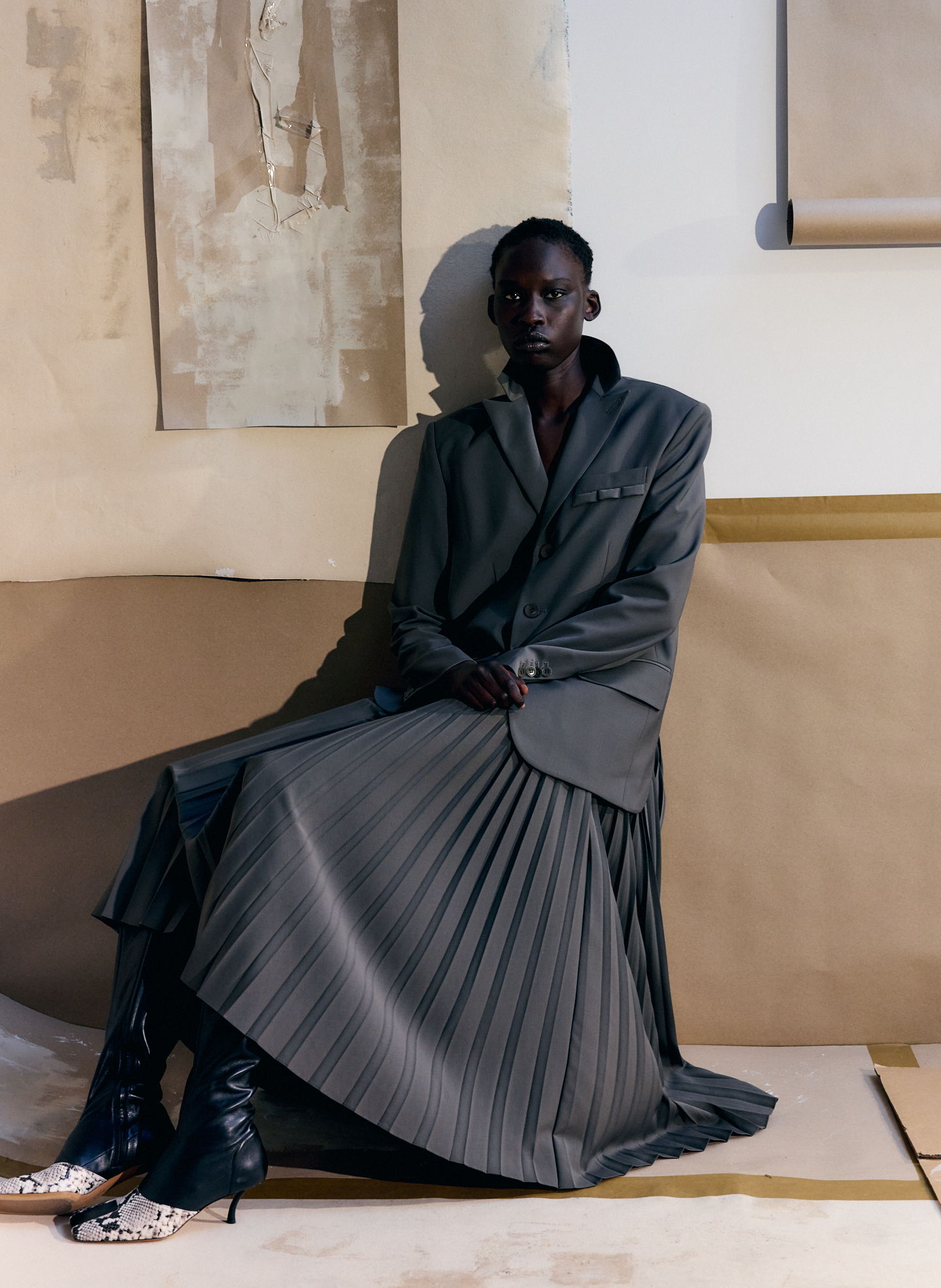 A person wearing a tailored blazer and pleated skirt, seated against a neutral backdrop.
