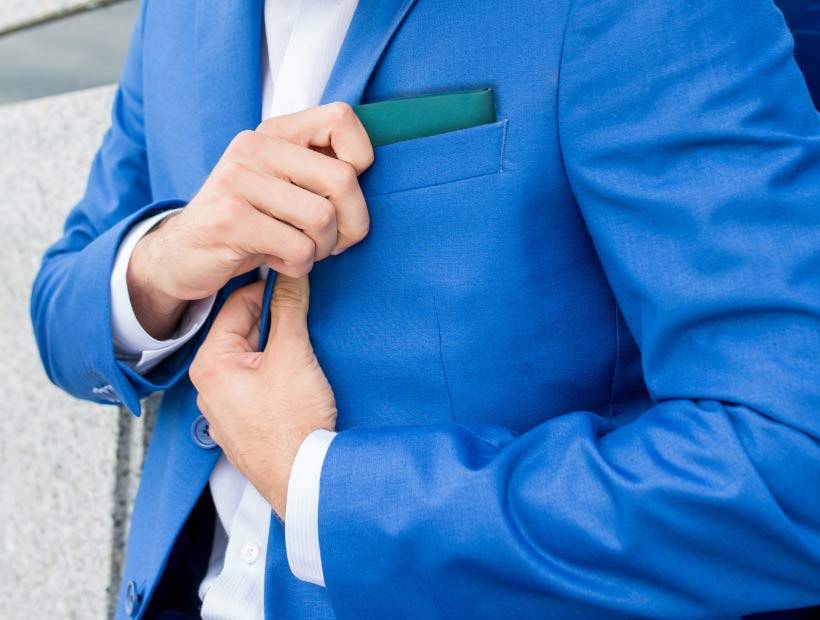 A man wearing a blue suit jacket and tucking in a blue/green pocket square 