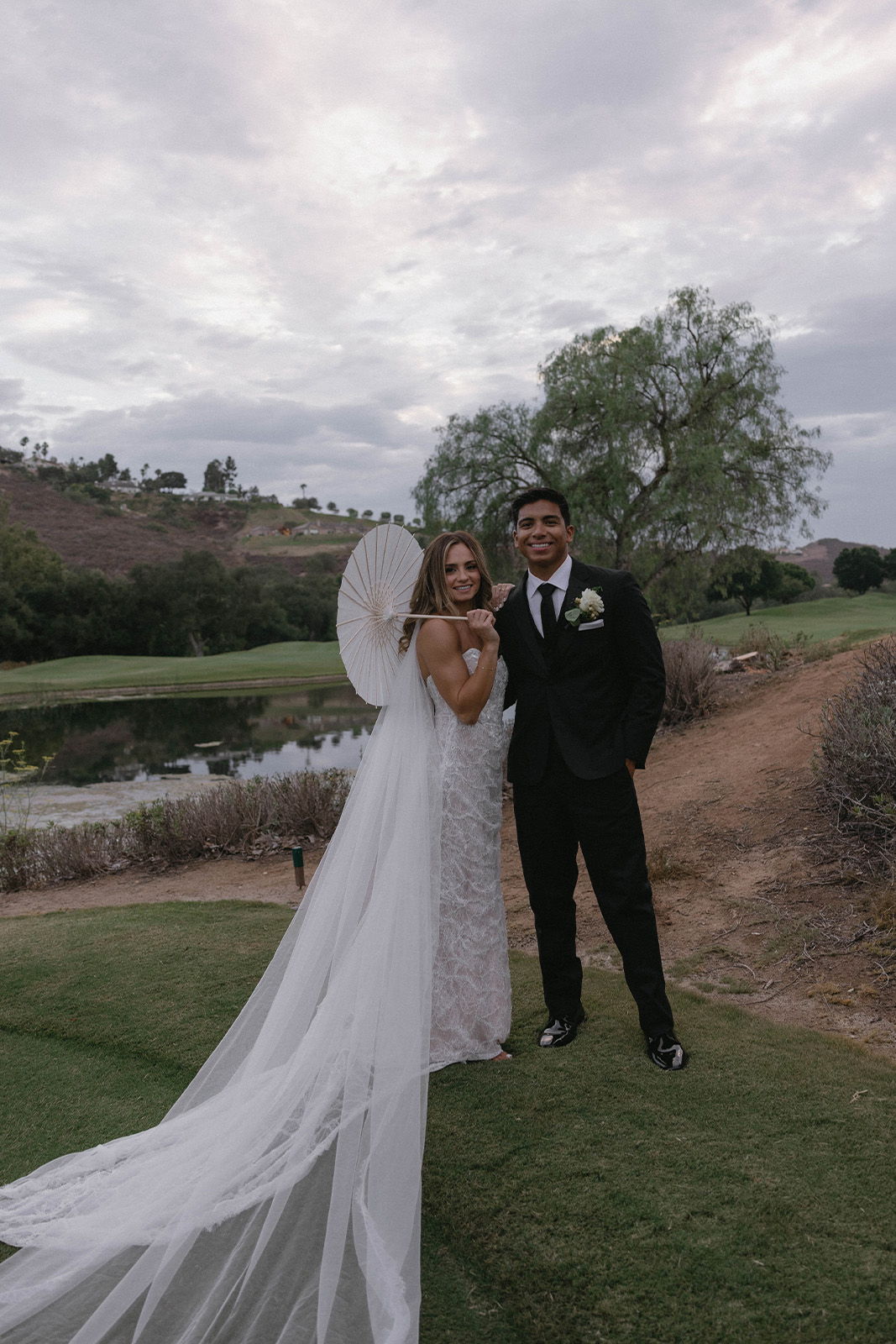 Bride and groom posing outdoors with a parasol and scenic background.
