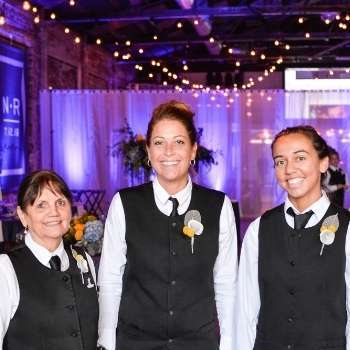 Three female caterers wearing black vests, white shirts and black ties at event