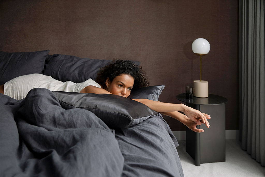 Model lays on bed styled with the Slate linen sheet set and duvet cover. The Line table lamp sits on a bedside table.  