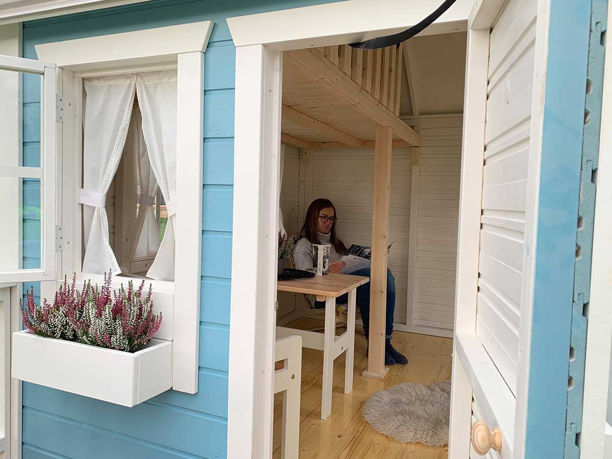 Mum reads a magazine in kids outdoor playhouse with opening windows and flower boxes by WholeWoodPlayhouses