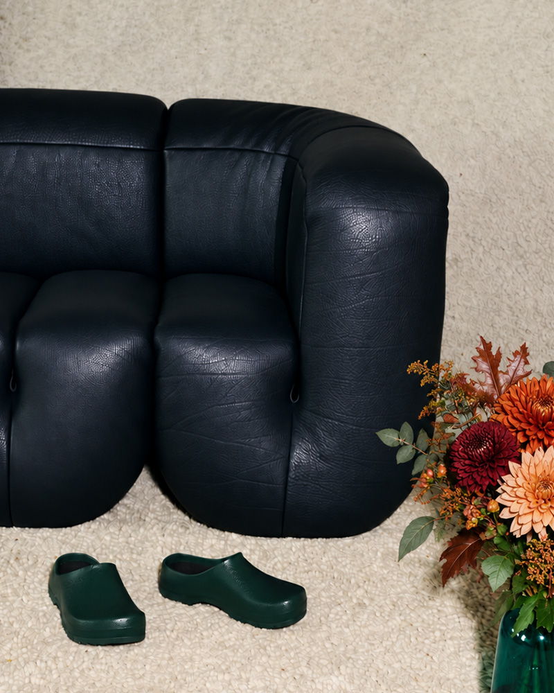 Black leather de Sede couch with green clogs and a flower arrangement on the floor.