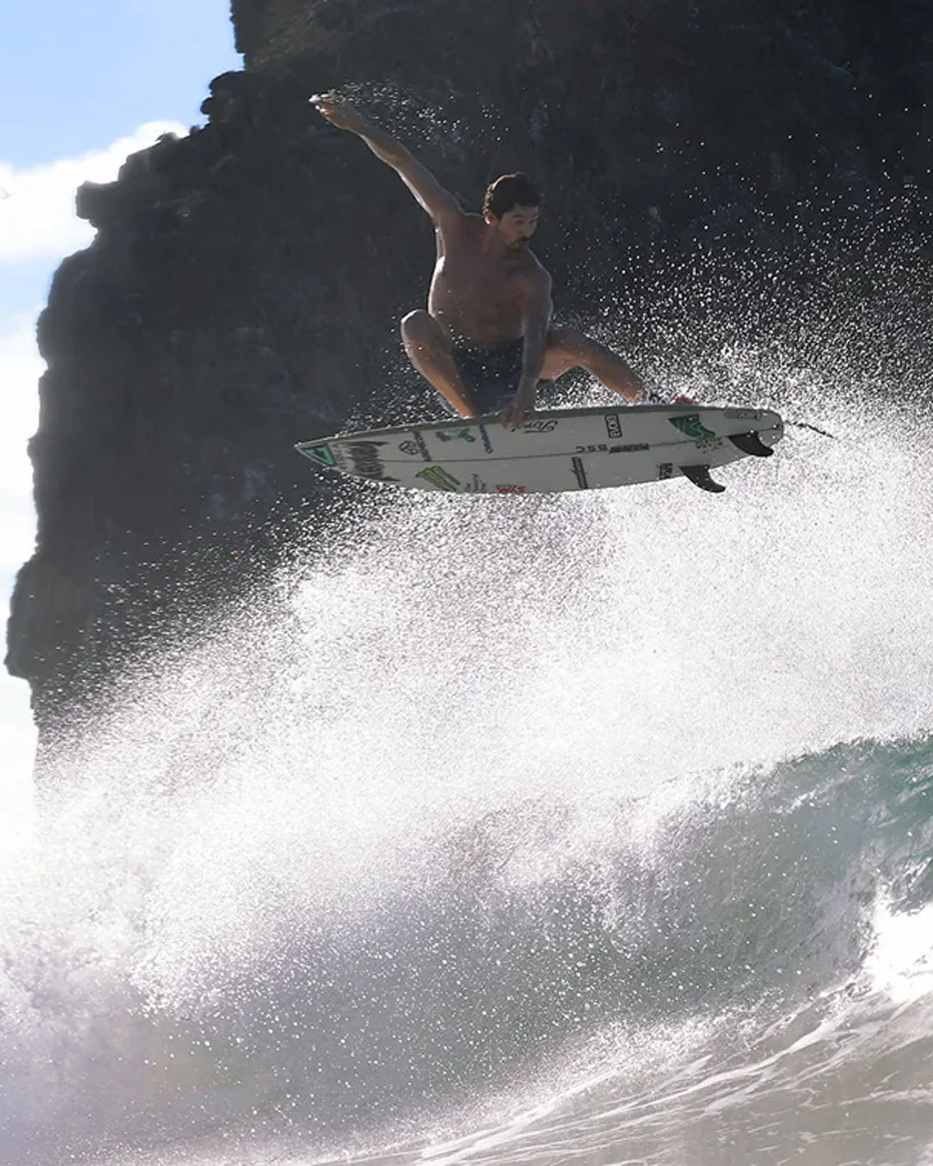 Surfer performing a jump over a wave with a rocky background.
