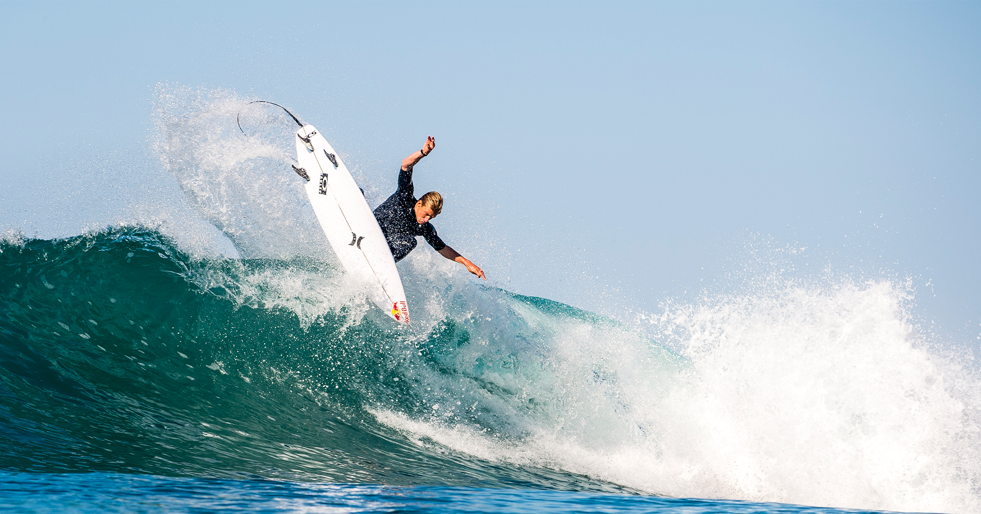 Surfer riding a wave, performing a maneuver against a clear blue sky.