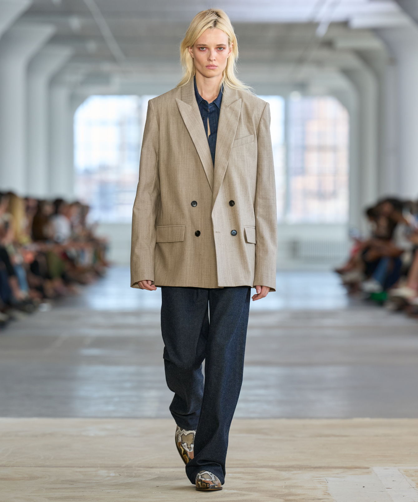Model walking down runway wearing oversized beige blazer and dark pants.