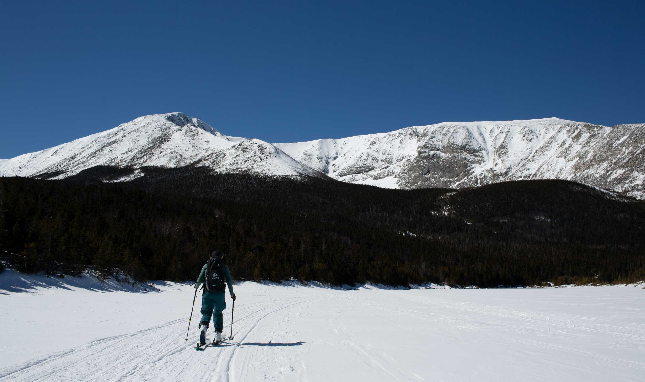 Person skiing on snow with mountains in the background under a clear blue sky.