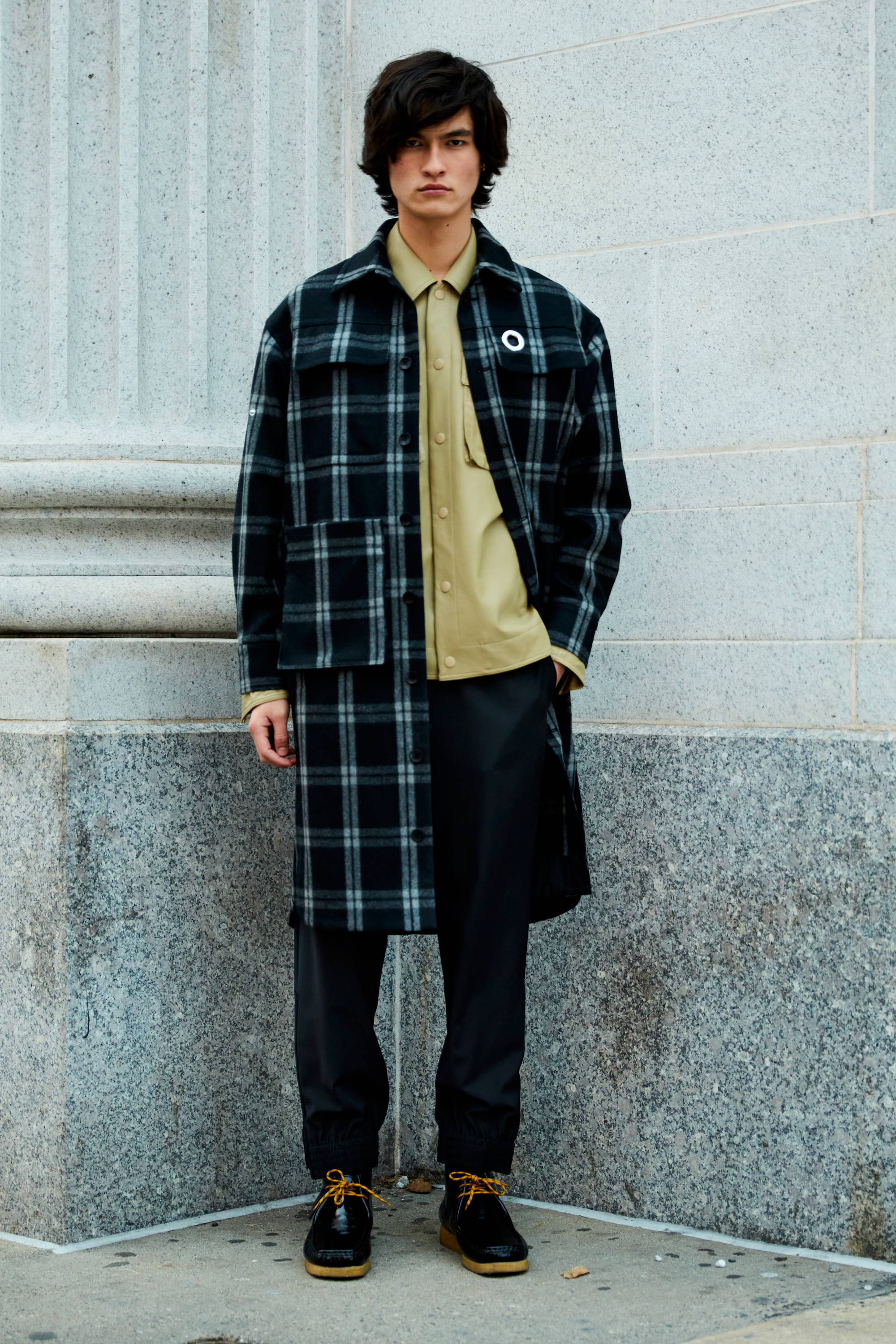 A person in a plaid coat and beige shirt stands against a stone wall.