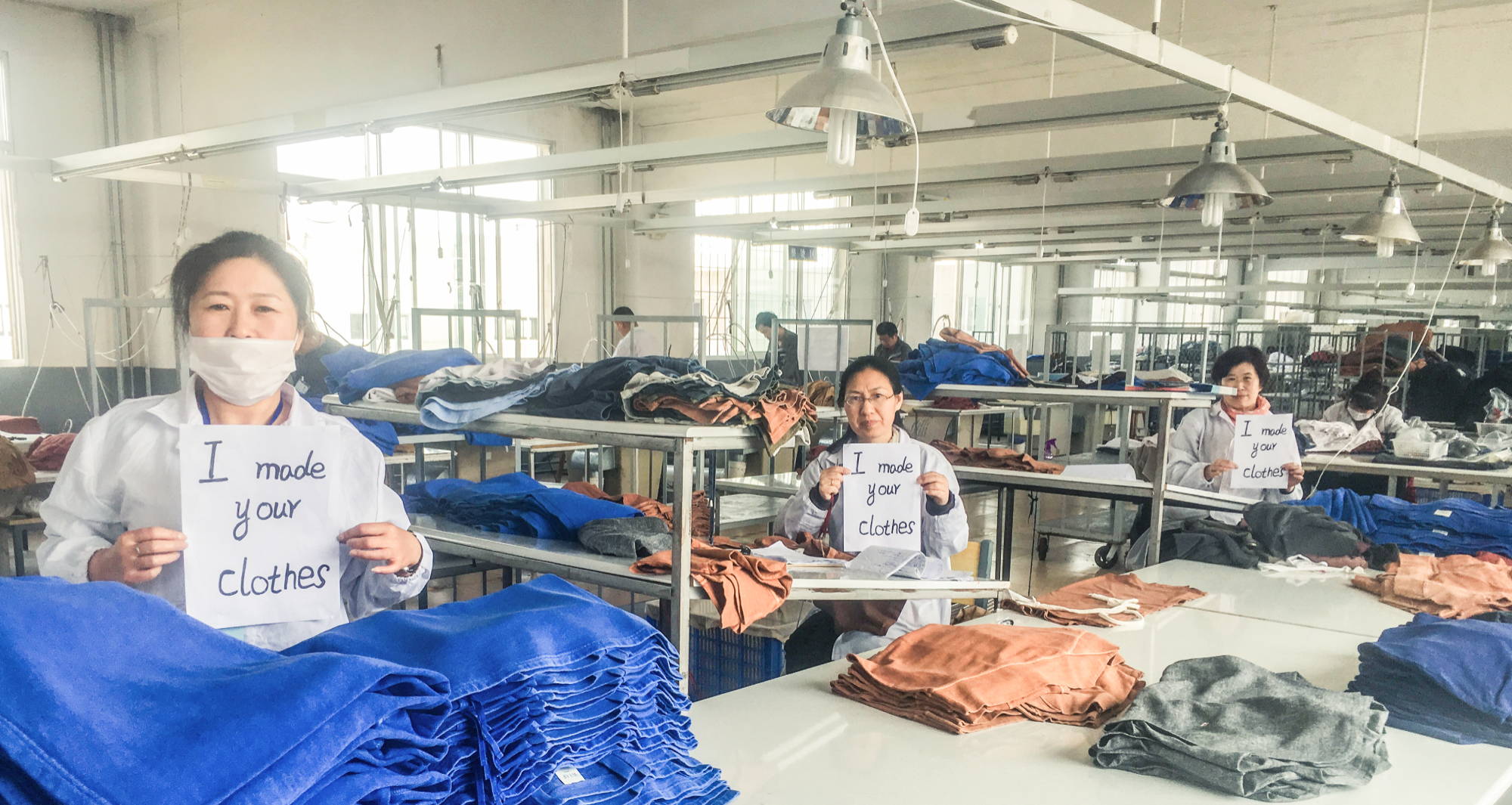  three women in white protective clothing in a clothing factory holding signs that say “I made your clothes”