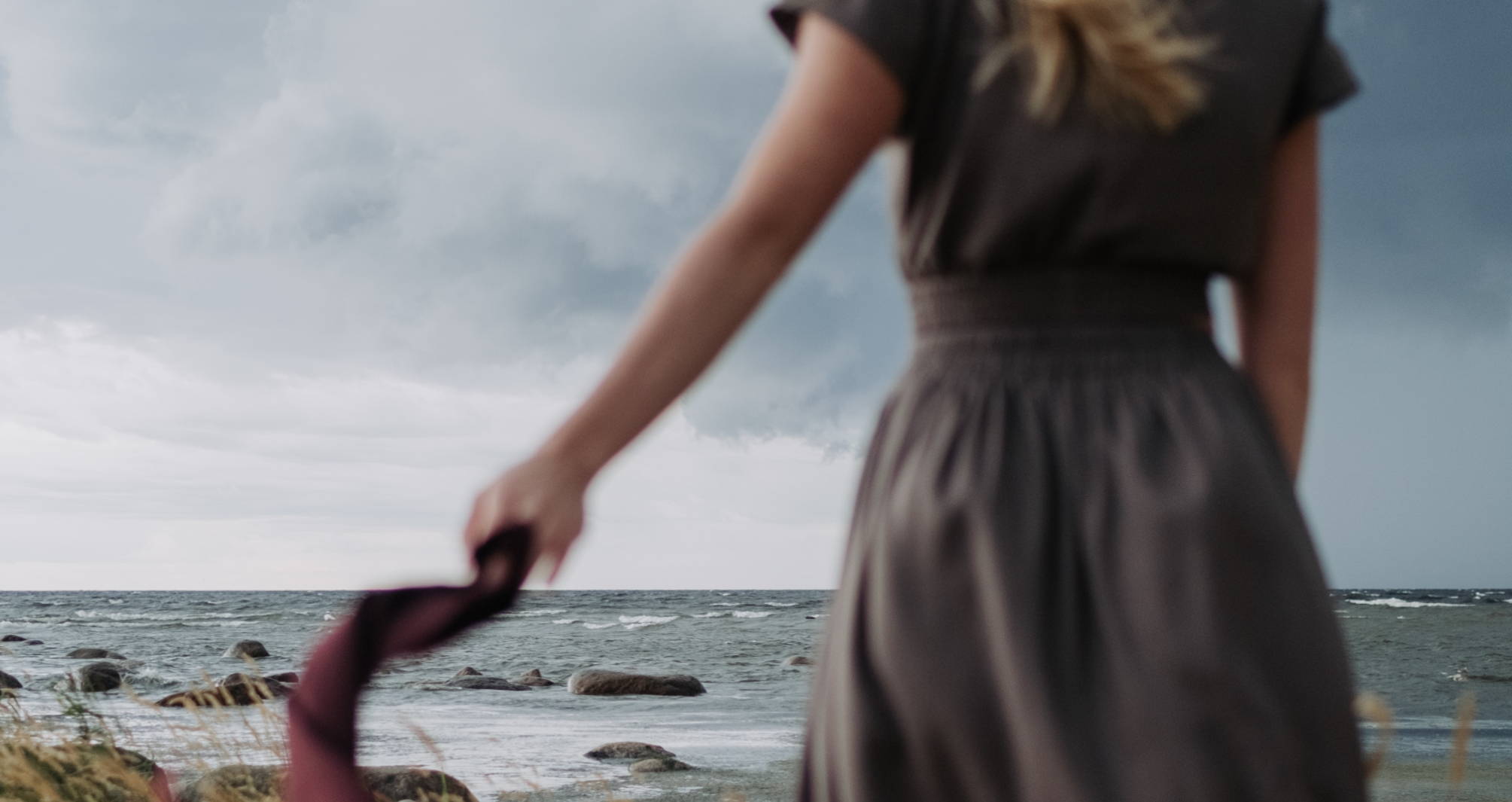 A woman in a gray linen dress waves a purple scarf, in soft focus, while the gray sky and ocean in front of her is in focus.