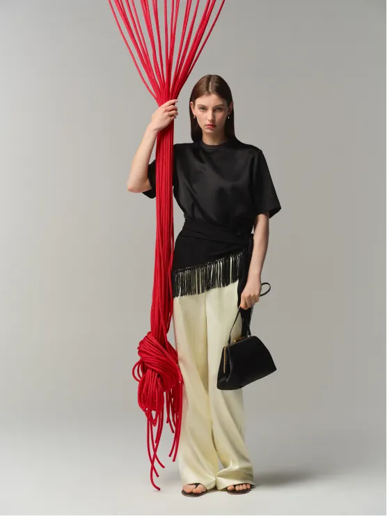Model holding red fabric, wearing black top and cream pants, with handbag.