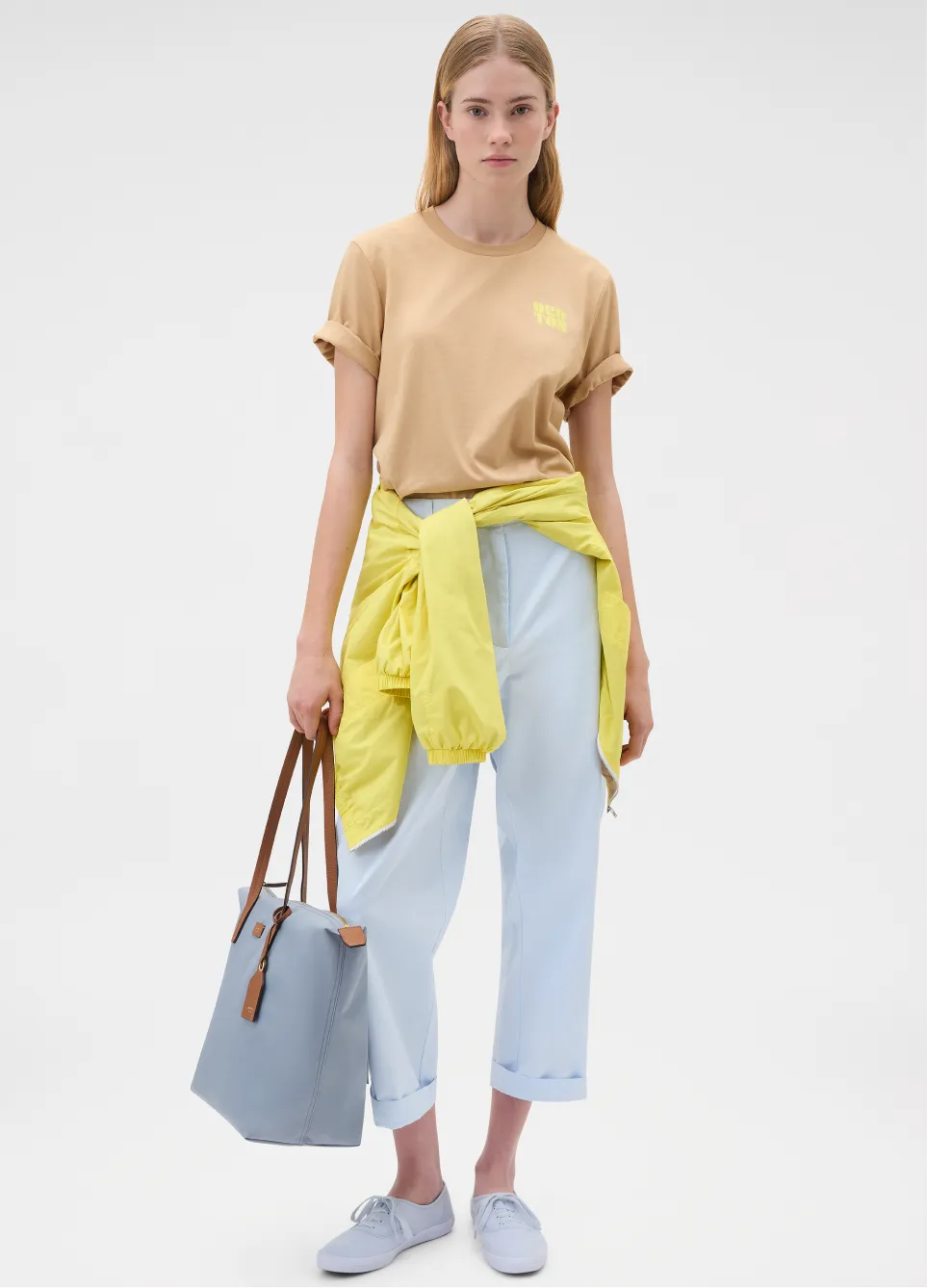 Model wearing a beige shirt and light blue pants, holding a tote bag.