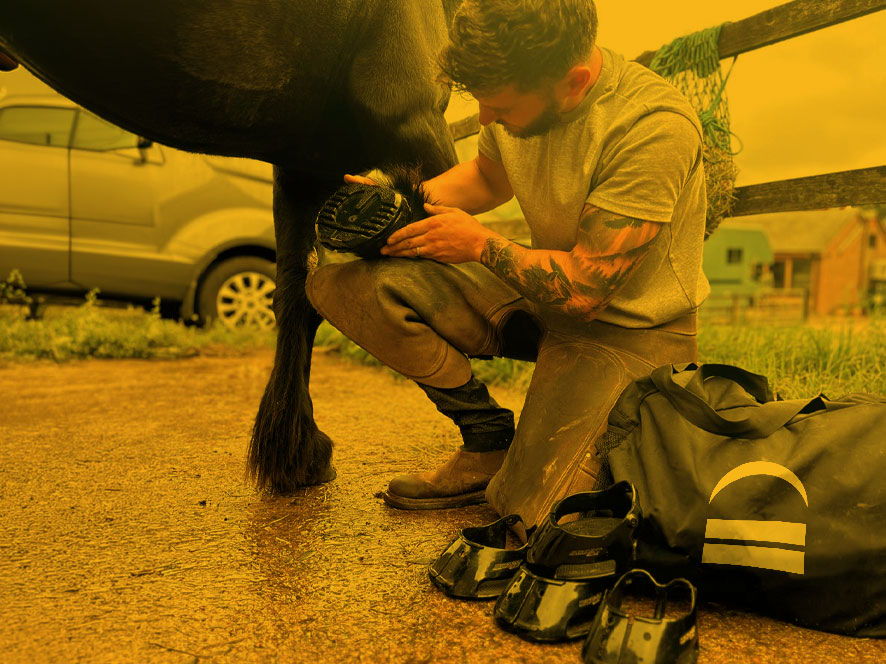 Person kneeling beside horse, adjusting equipment on a dirt path.