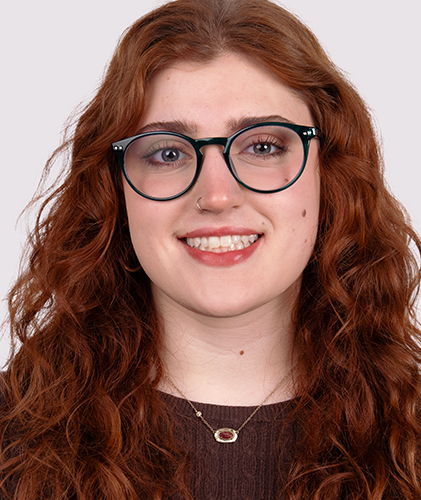 A smiling person with curly red hair and glasses against a plain background.