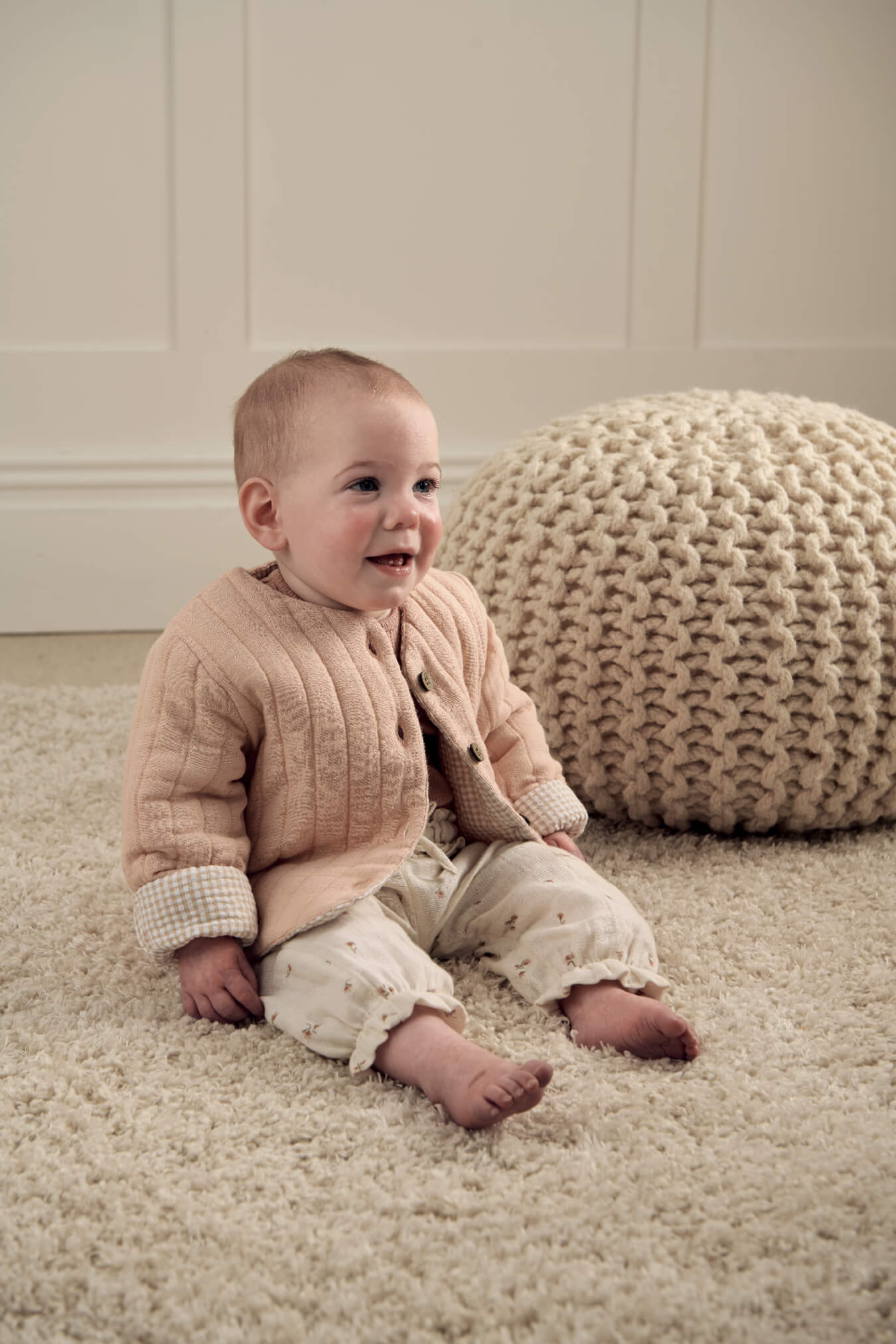 Baby sitting on a soft rug, wearing a WTTW Reversible Quilted Jacket and smiling.