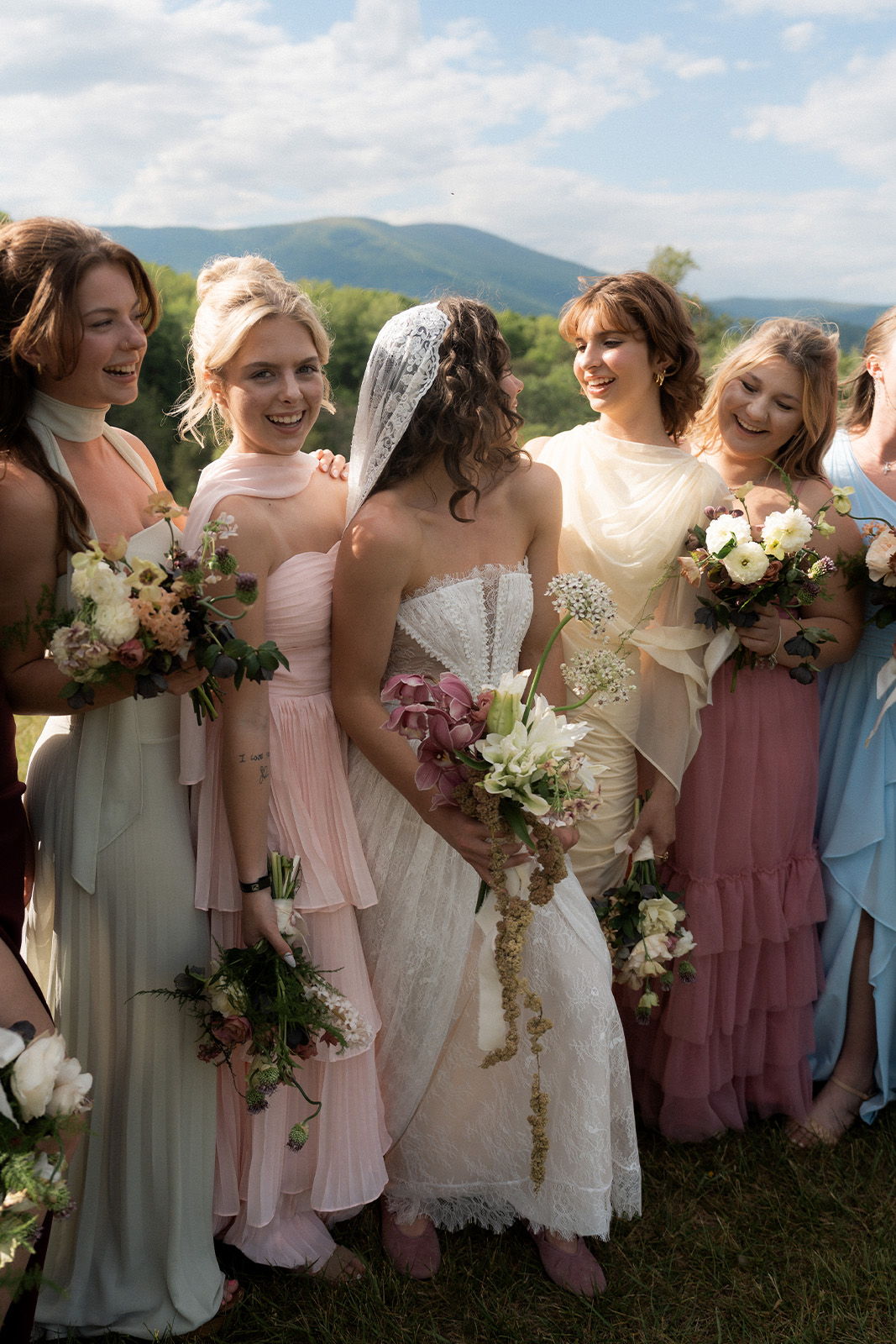 Bridal party in pastel dresses holding bouquets, outdoors with mountains in background.
