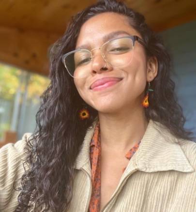 Woman with big nose wearing Enchanted Gold, Cat-Eye Eyeglasses in Gold Metal with a printed shirt, sunflower earrings and a beige jacket
