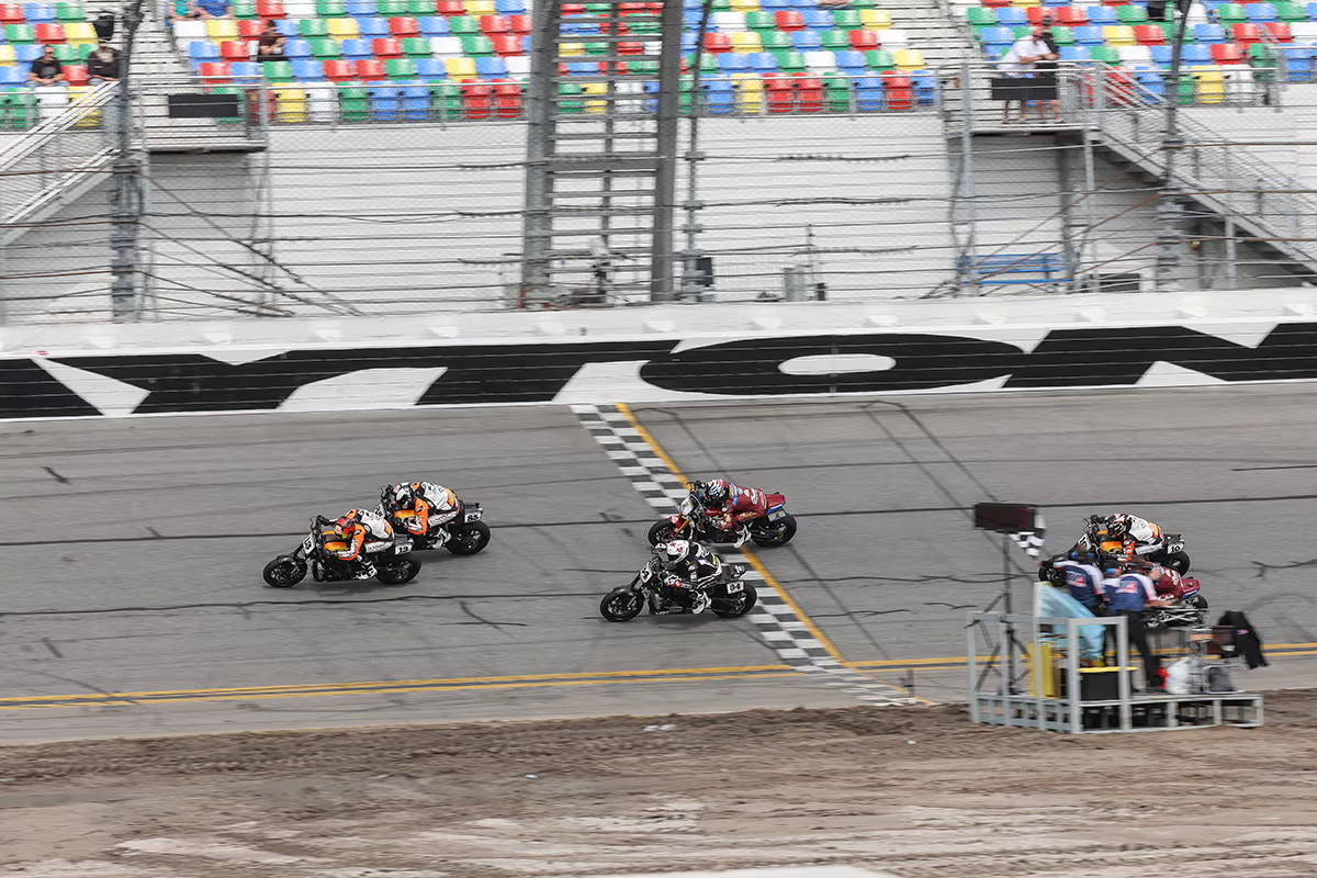 Multiple motorcycle racers approaching the finish line at Daytona International Speedway, the checkered flag waved by an official.
