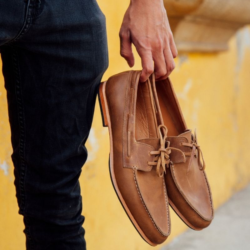 A close-up lifestyle photograph of a person holding a pair of tan or caramel brown leather Adelante Nautico boat shoes by the heels in one hand. The person is wearing dark textured pants or jeans, standing next to a weathered, vibrant yellow stucco wall in bright daylight. The leather boat shoes feature classic matching laces and traditional moc-toe stitching.