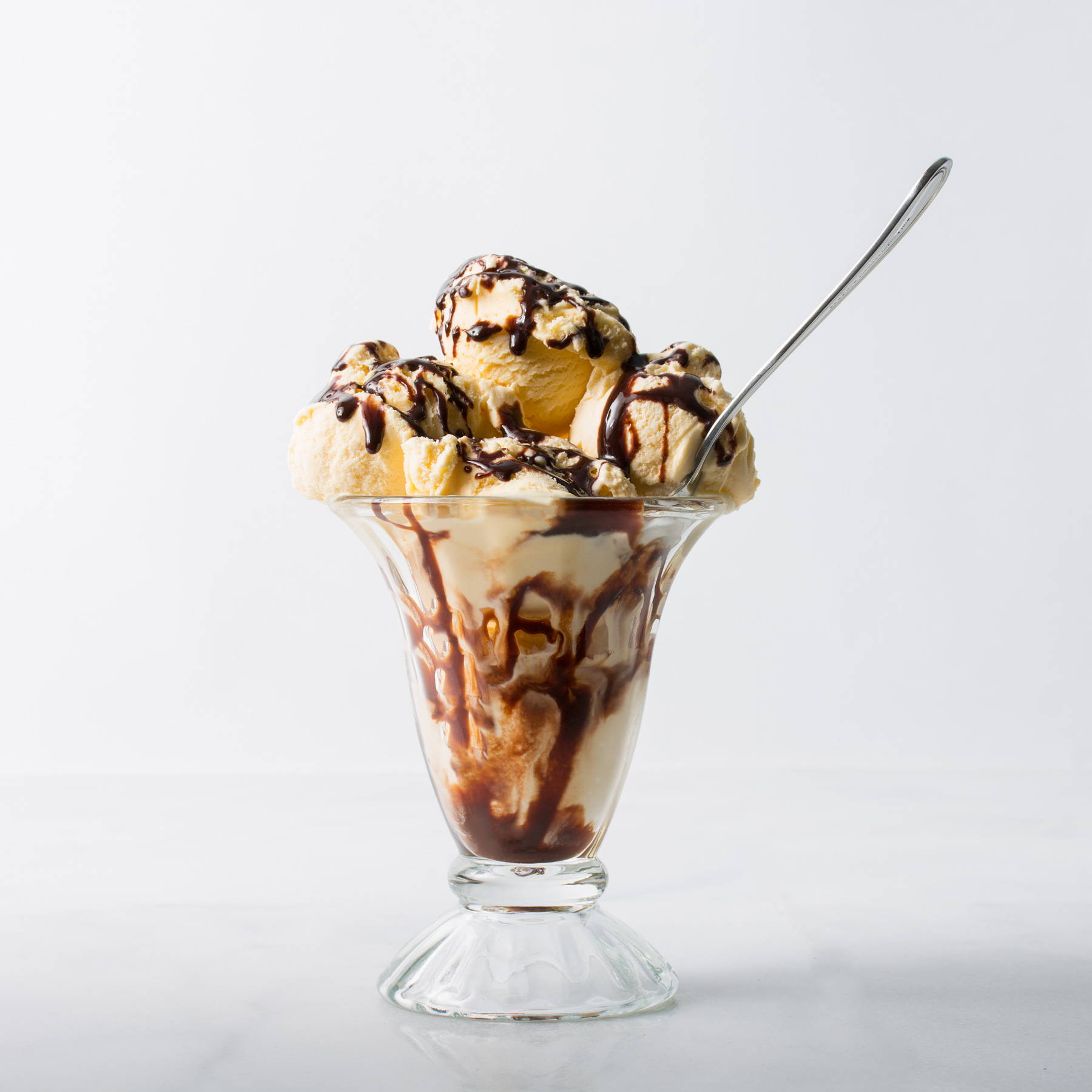 Hot caramel fudge sauce drizzled over scoops of vanilla ice cream in a glass sundae bowl with a spoon.