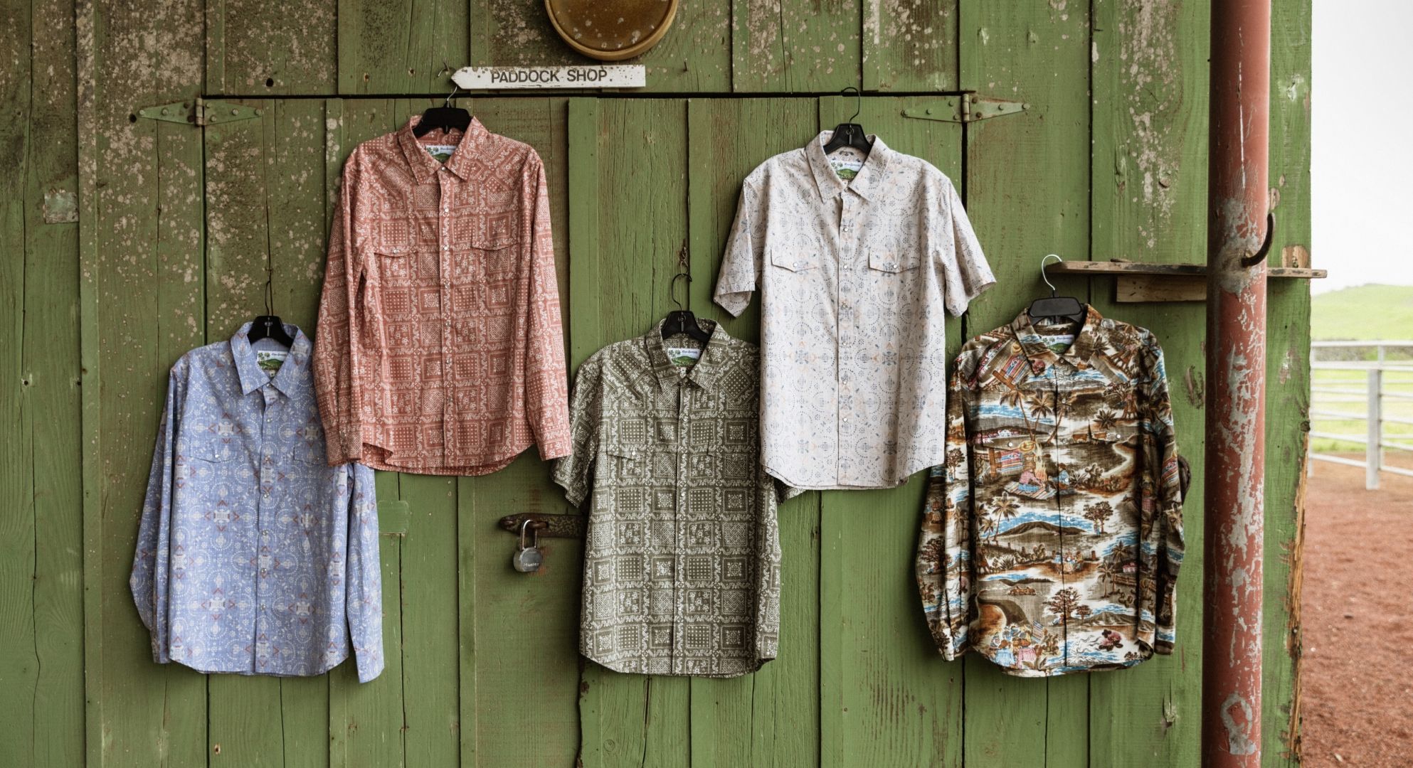 Five shirts hanging on hooks against a green barn wall.