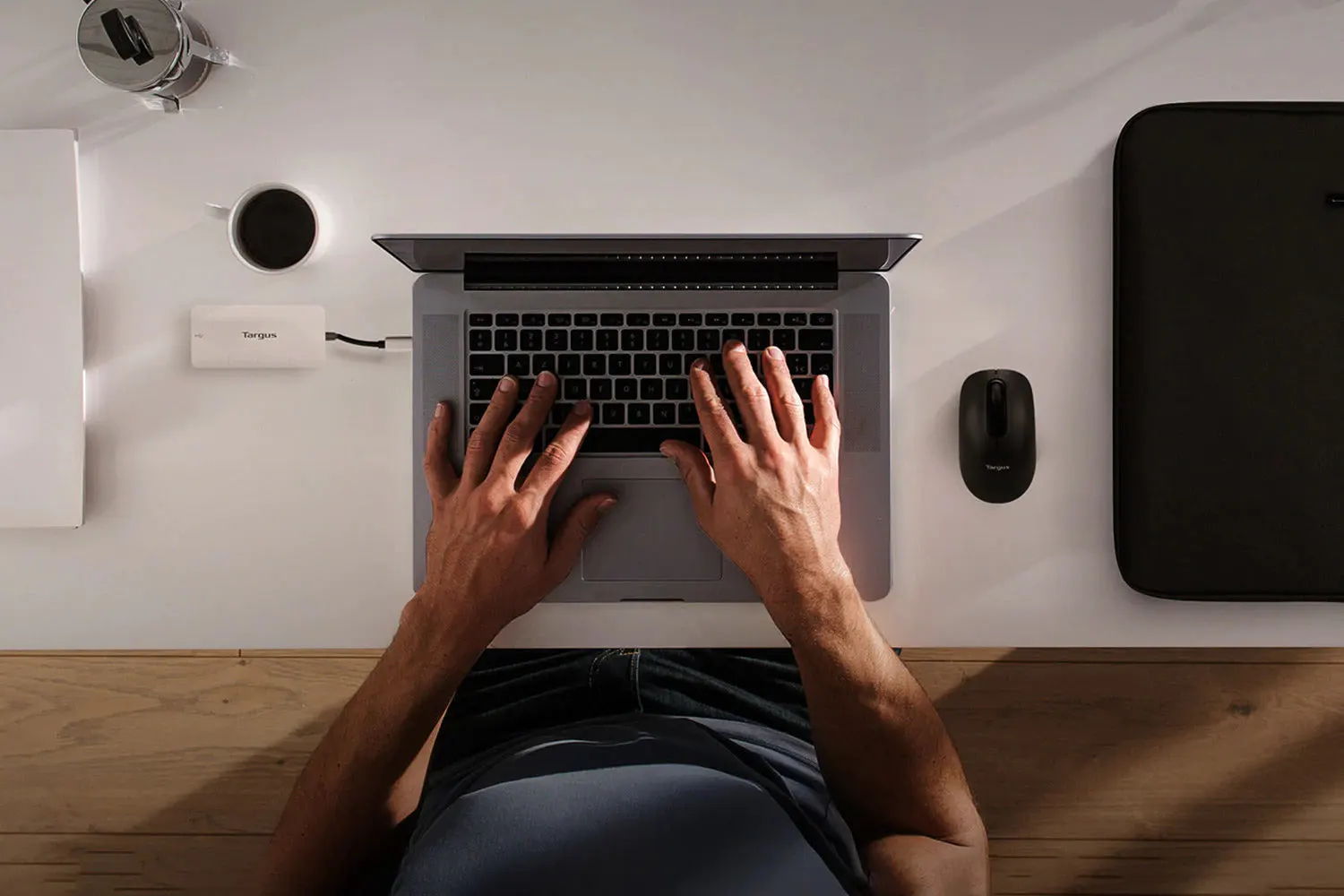Overhead shot of hands typing on a laptop at a desk with coffee, a power bank, mouse, and laptop sleeve.