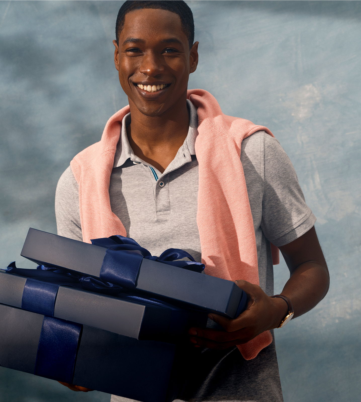 Smiling young man holding gifts with a pink sweater draped over his shoulders.