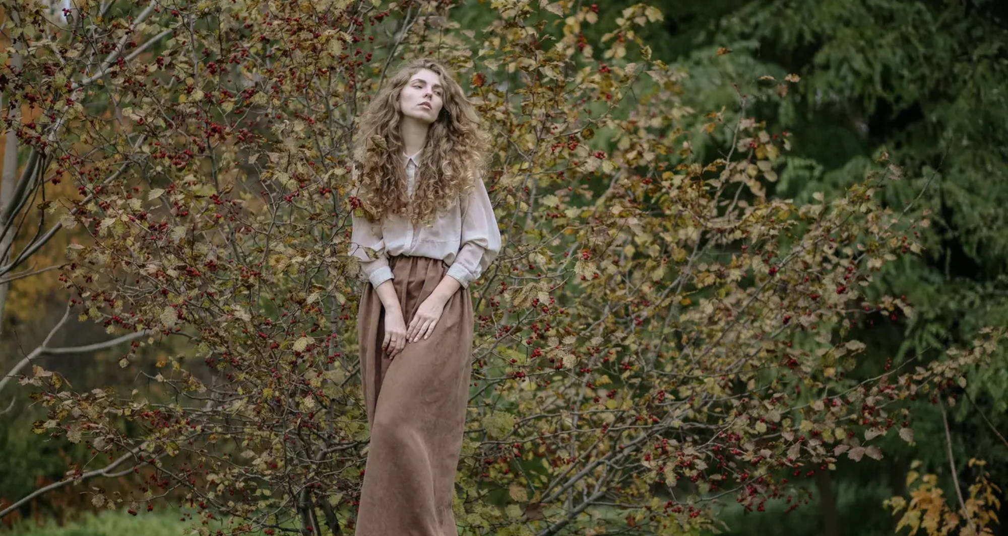 A woman with long curly hair wearing a white blouse and brown skirt stands on a tree stump in a wooded area.