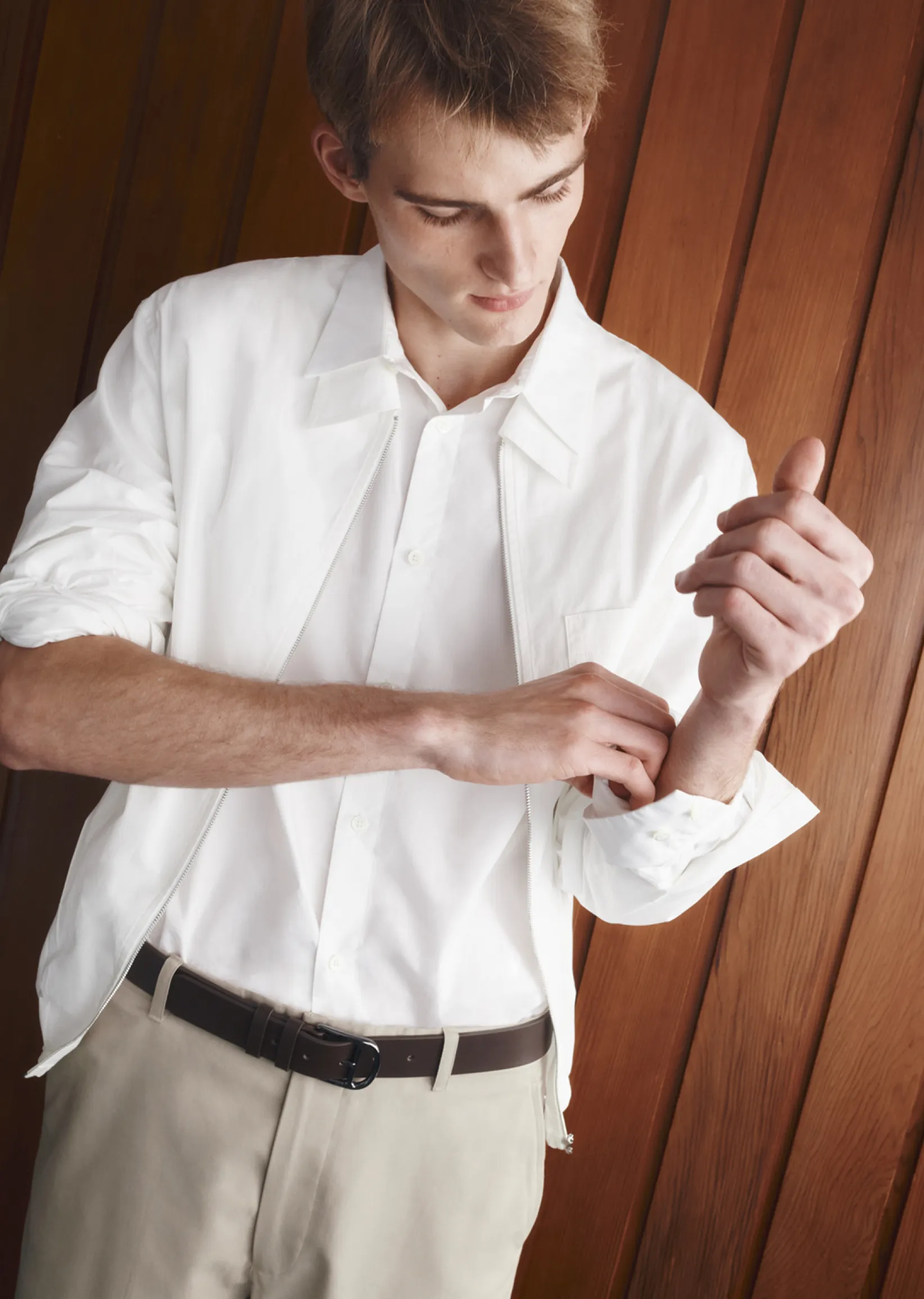 A young man adjusts his shirt cuff against a wooden background.