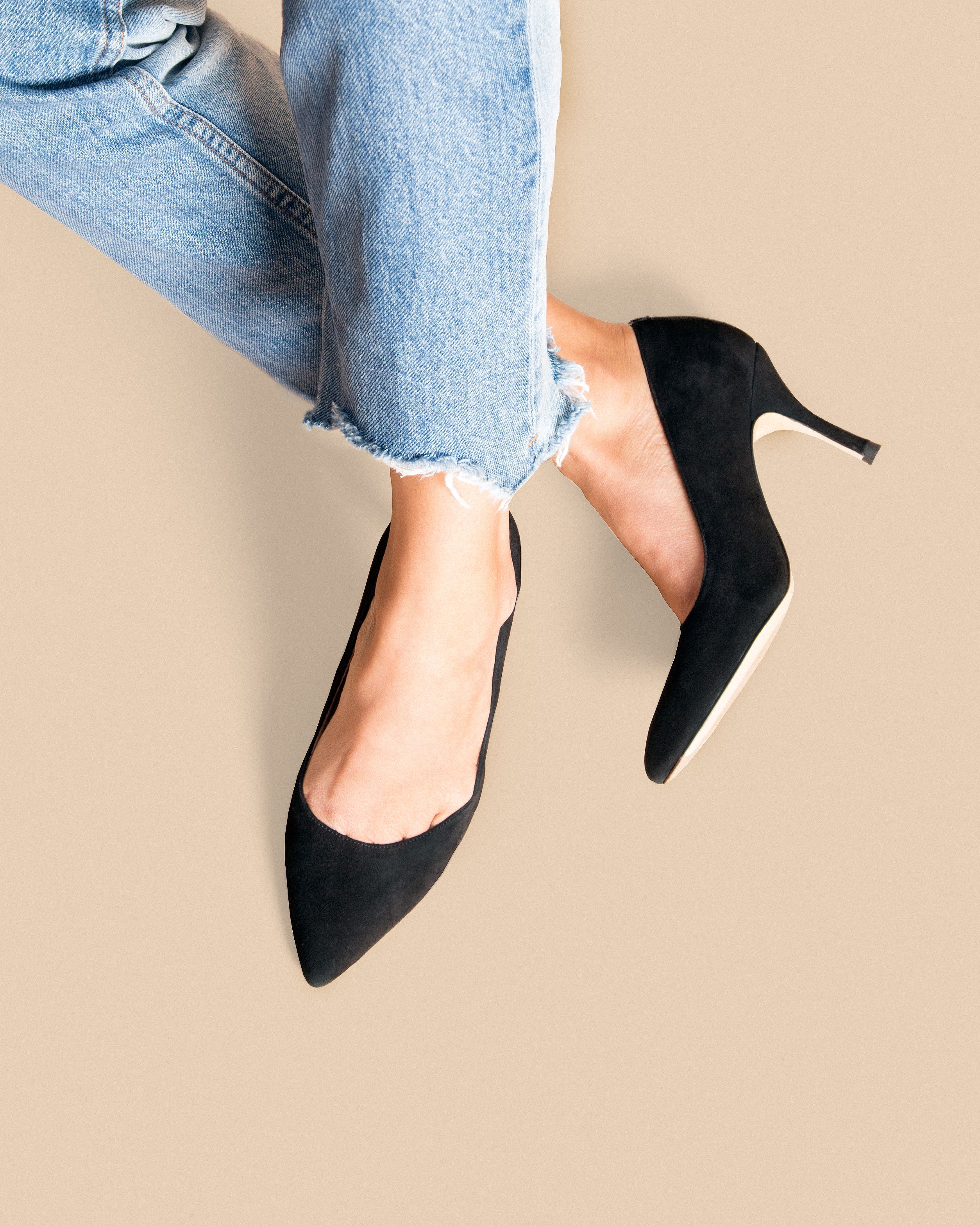 Black high heels worn with distressed jeans on a beige background.