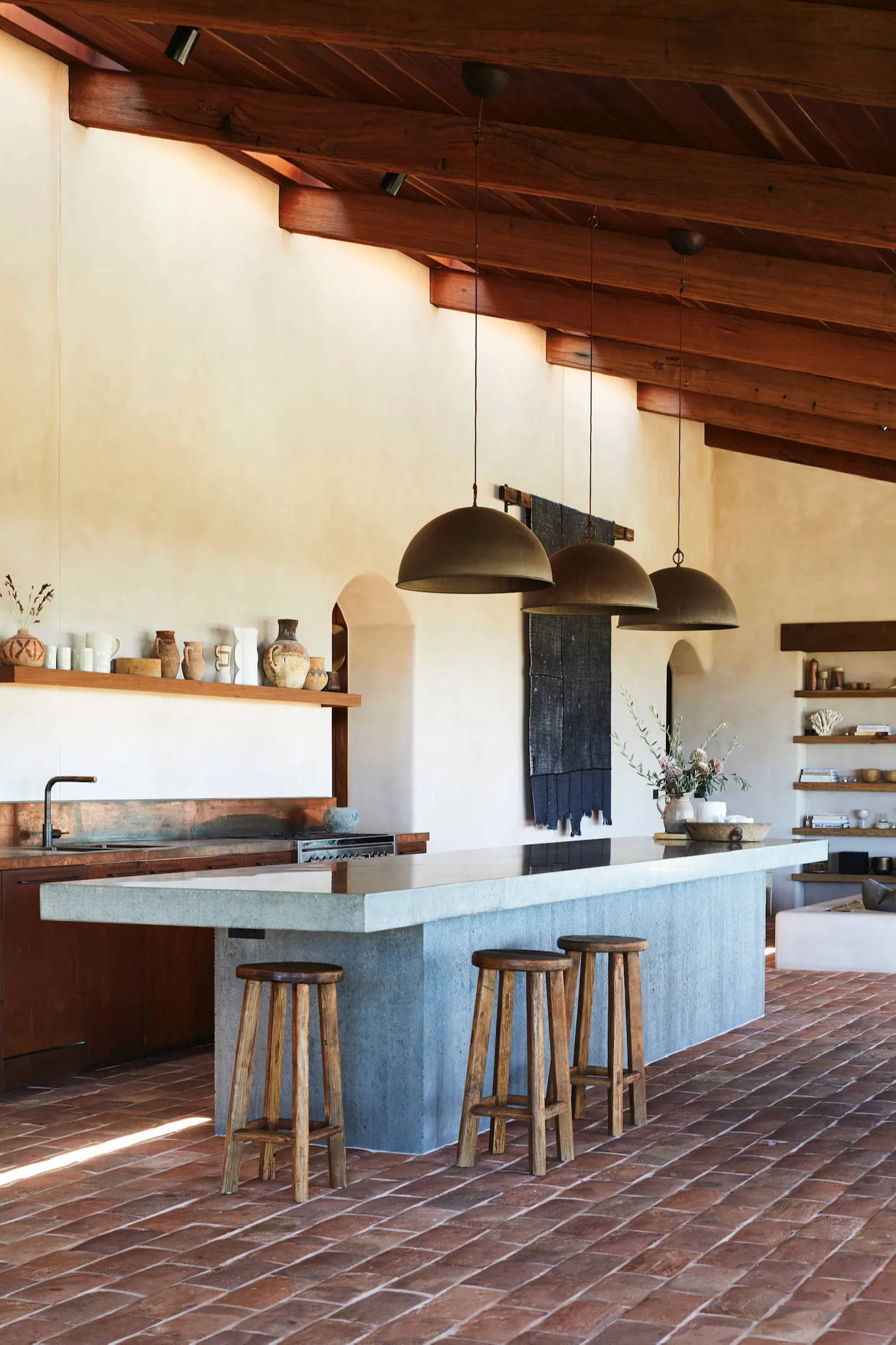 Outdoor kitchen with concrete island, wooden stools, and metal pendant lights.