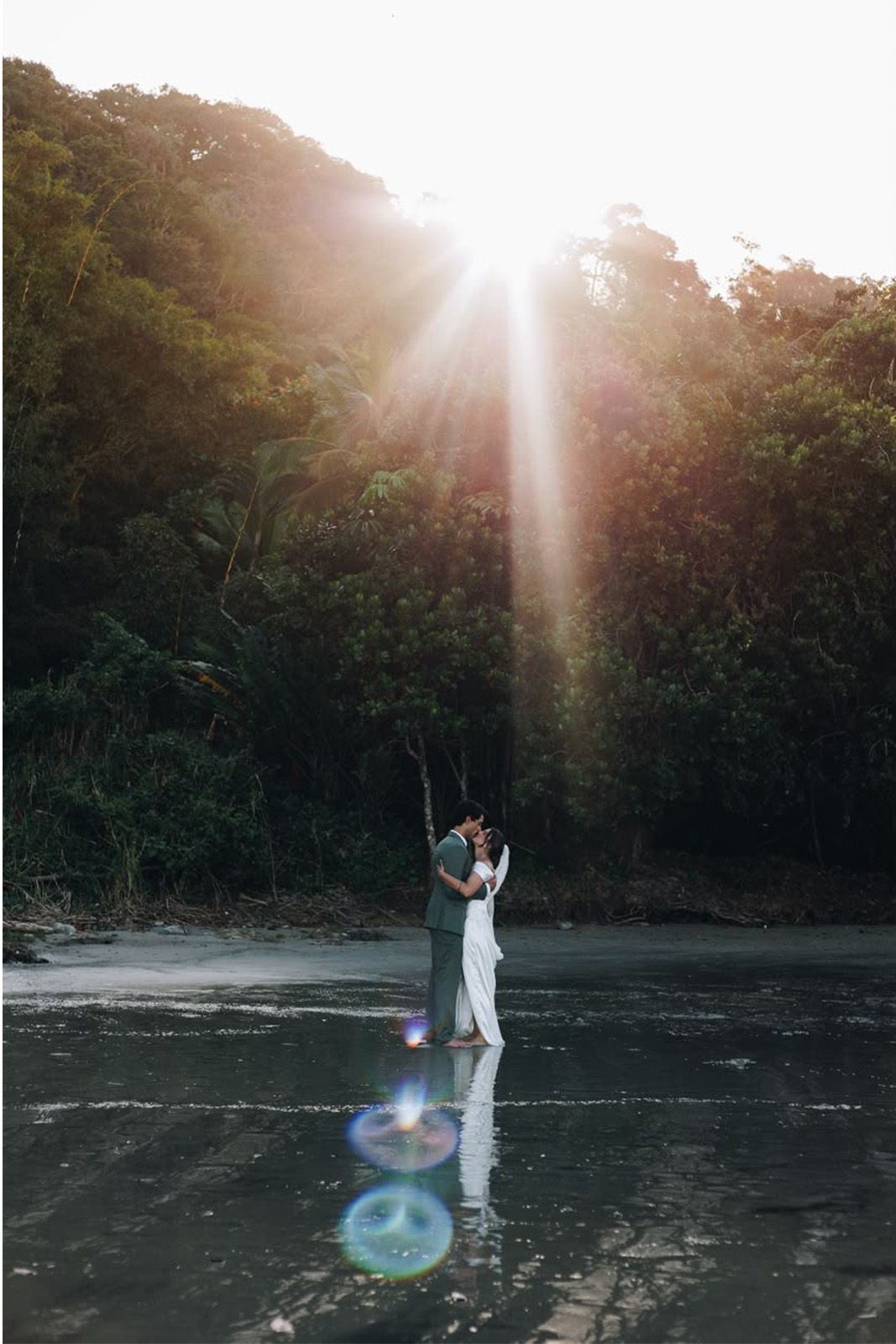 Couple embracing on a beach at sunset, surrounded by lush greenery.