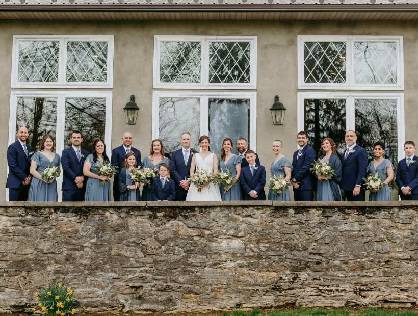 A large wedding party in blue, posing outside near a brick wall