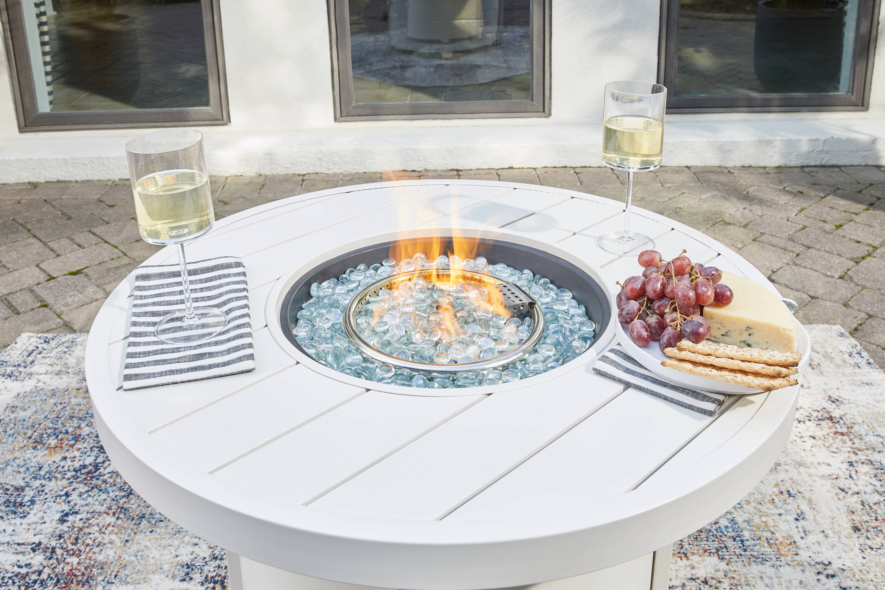 A white fire pit tables holds two champagne glasses and a bowl of grapes.