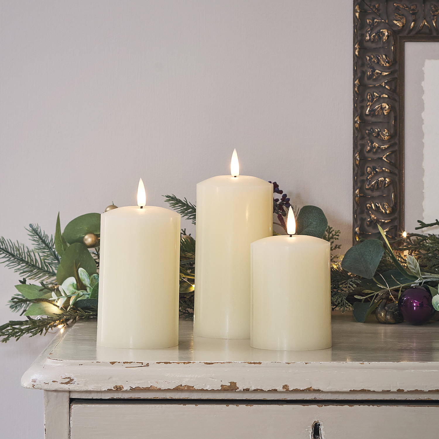 3 ivory TruGlow LED candles on a sidetable with a Christmas garland in the background.