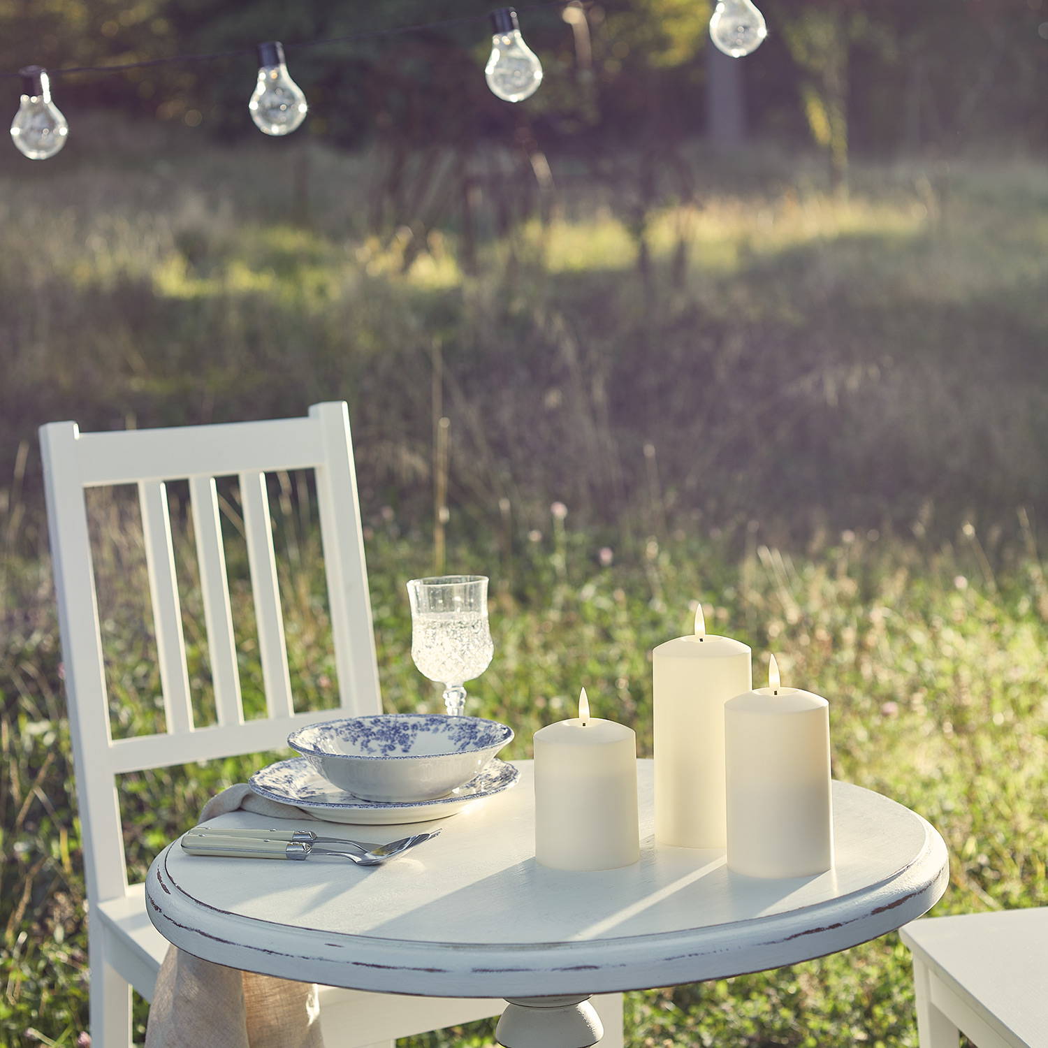 An outdoor table with 3 ivory LED candles and festoon lights hanging above.