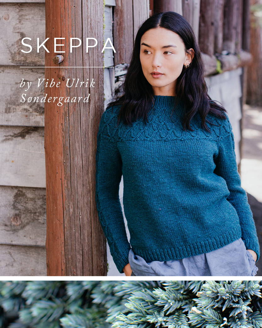 A woman leans against the outside wall of a log cabin with her hands in her pockets. She models a hand knit sweater with the title SKEPPA by Vibe Ulrik Sondergaard
