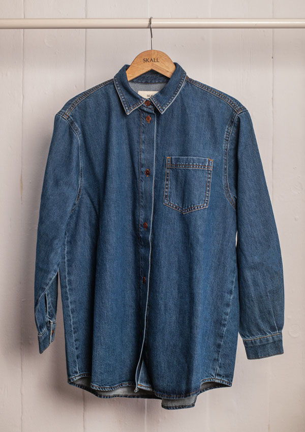 Denim shirt hanging on a wooden hanger against a white wall.