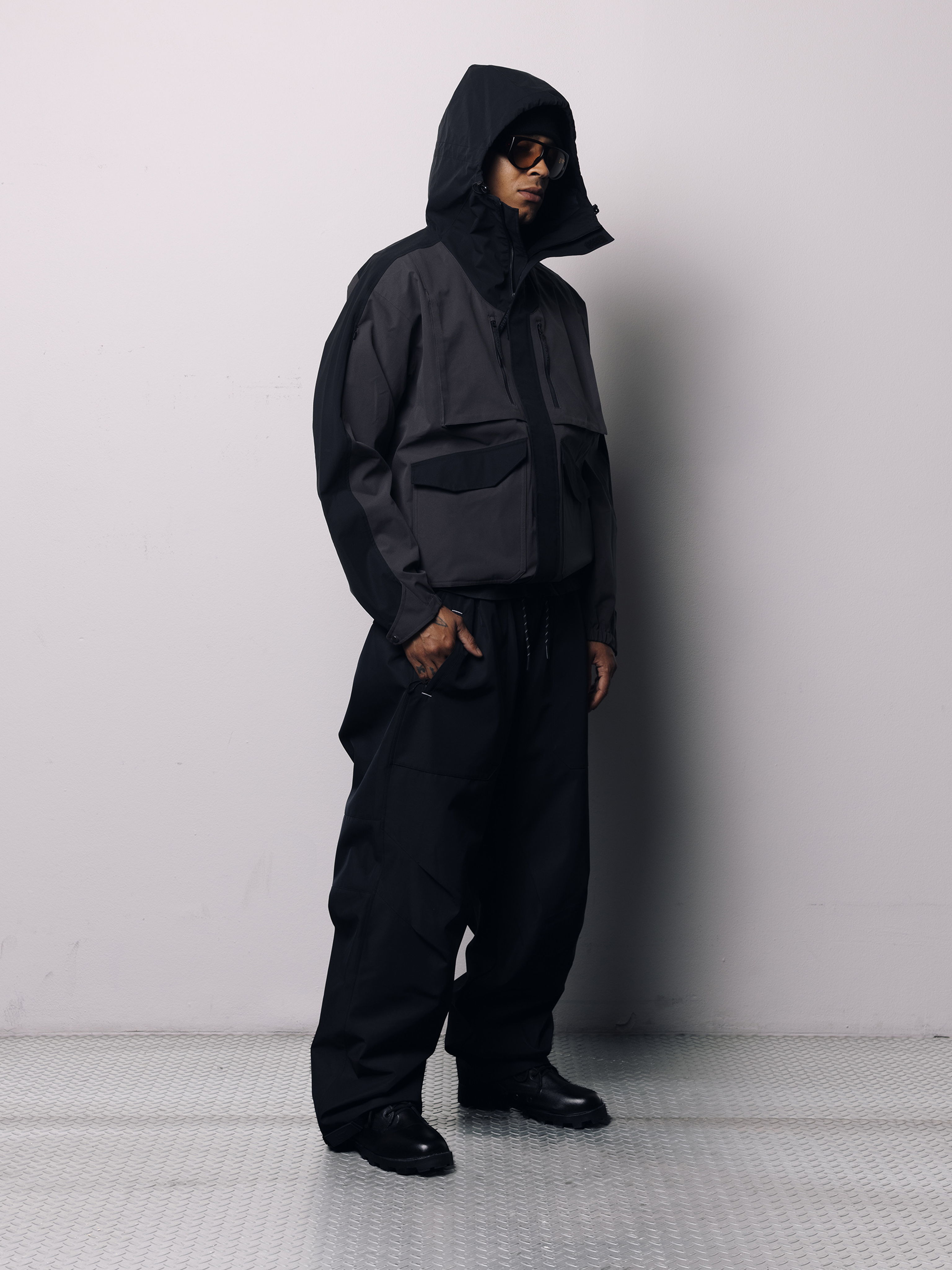 Person wearing a stylish all-black outfit with a hood, standing against a plain background.