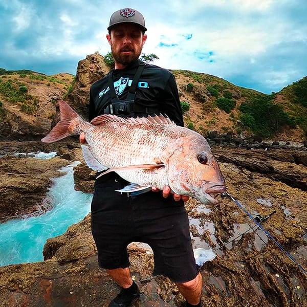 Blake Foster next to a river, holding up a large fish to the camera, while wearing an SA Company hooded performance shirt and a hat.