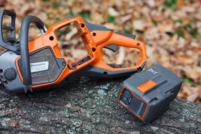 Orange chainsaw and battery pack on a log with fallen leaves.