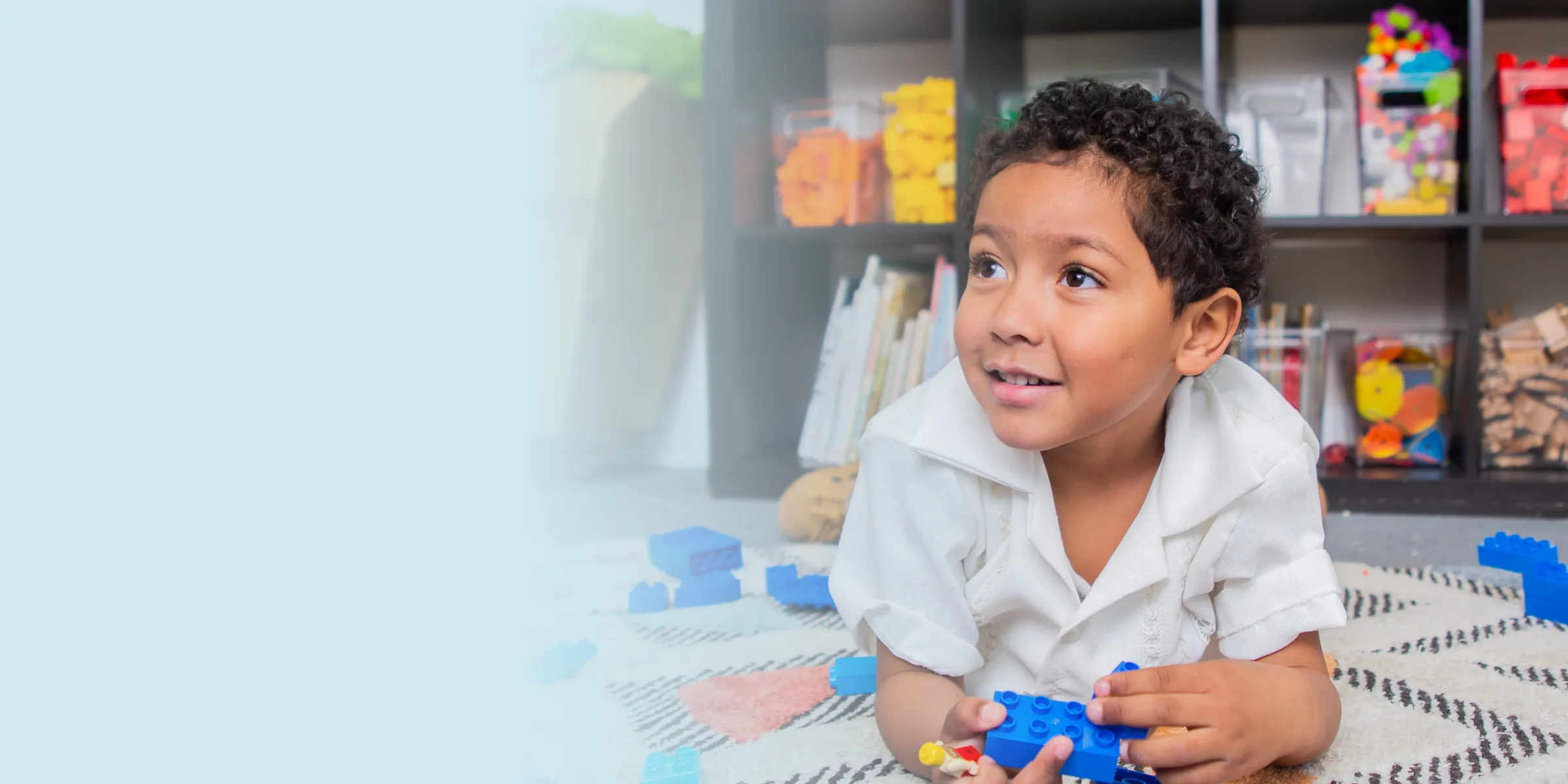 A child with curly hair is playing on a playmat in a nursery. He looks up and to the left slightly, with an expression of hope on his face.
