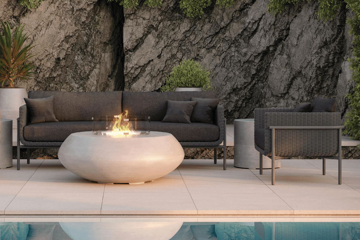 Modern outdoor lounge with dark woven sofa and chair facing a low, oval concrete fire pit with flames beside a swimming pool and rock wall.