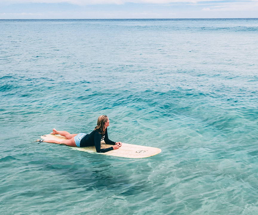 Photo of a female laying on her stomach on a surfboard in the water. There are no waves.