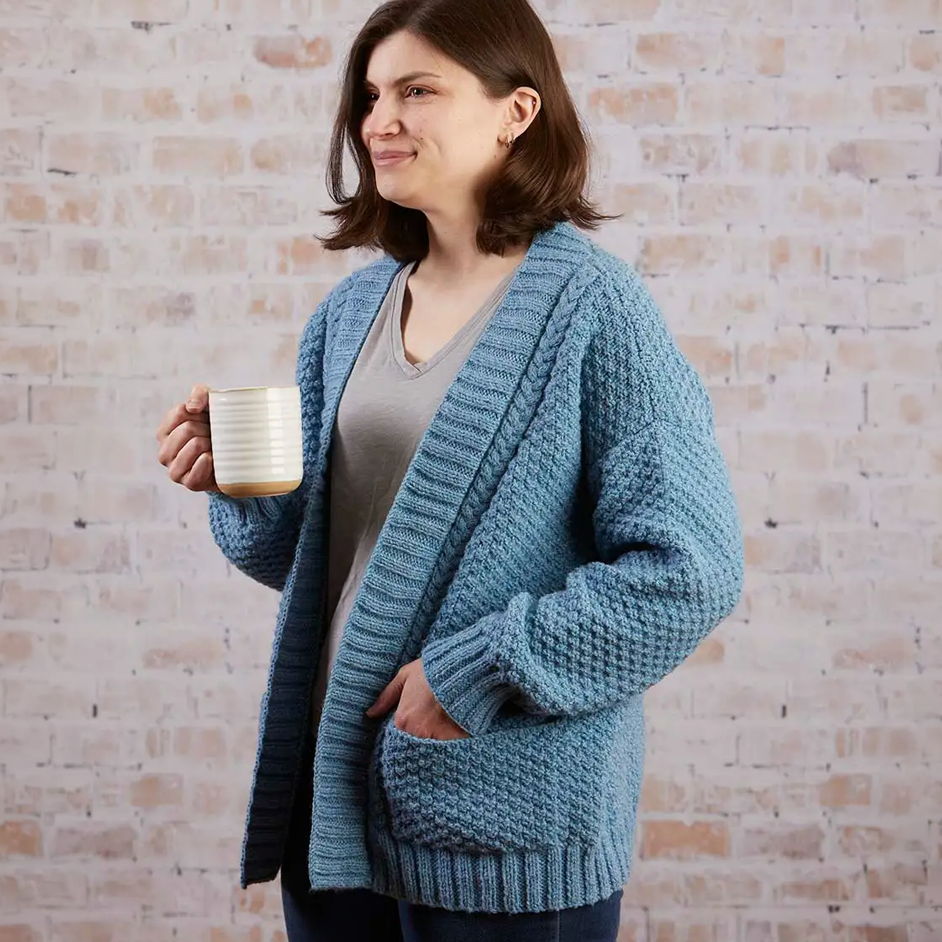 Woman in a blue cardigan holding a mug, standing against a brick wall.