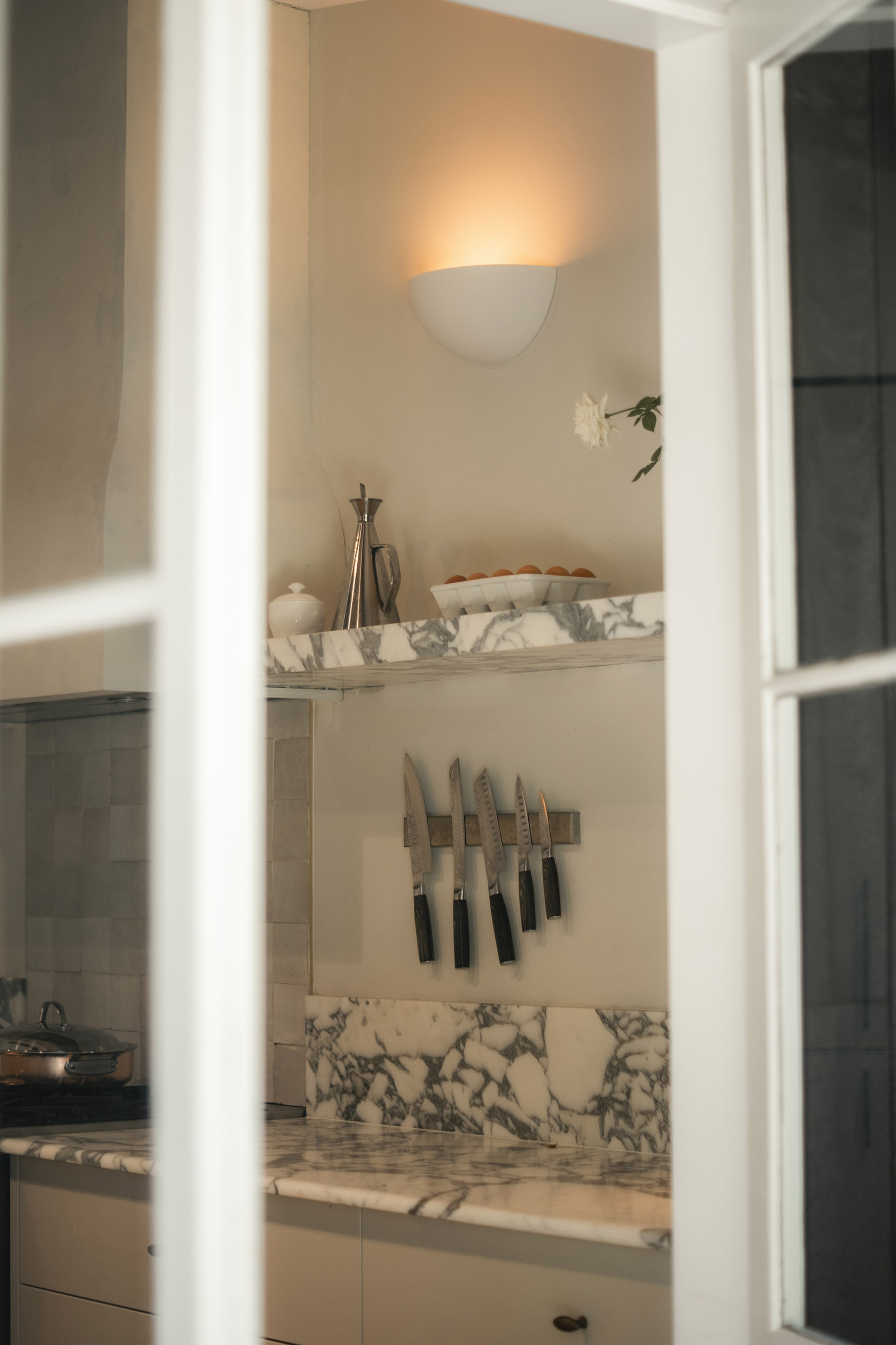 A modern kitchen through a window, featuring marble countertops and a plaster wall light