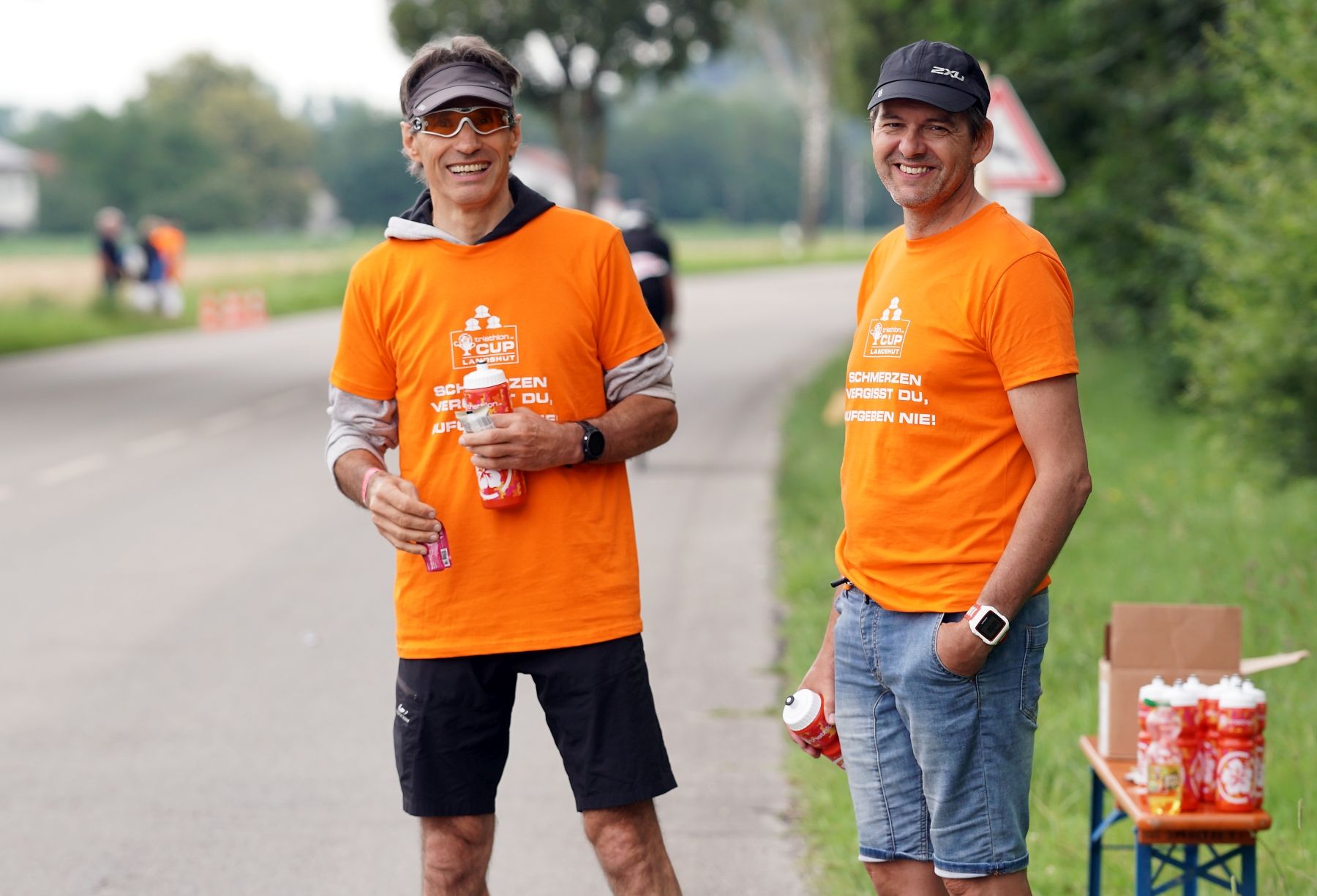 Zwei Männer in orange-farbenen T-Shirts reichen Radfahrern am Straßenrand Trinkflaschen an