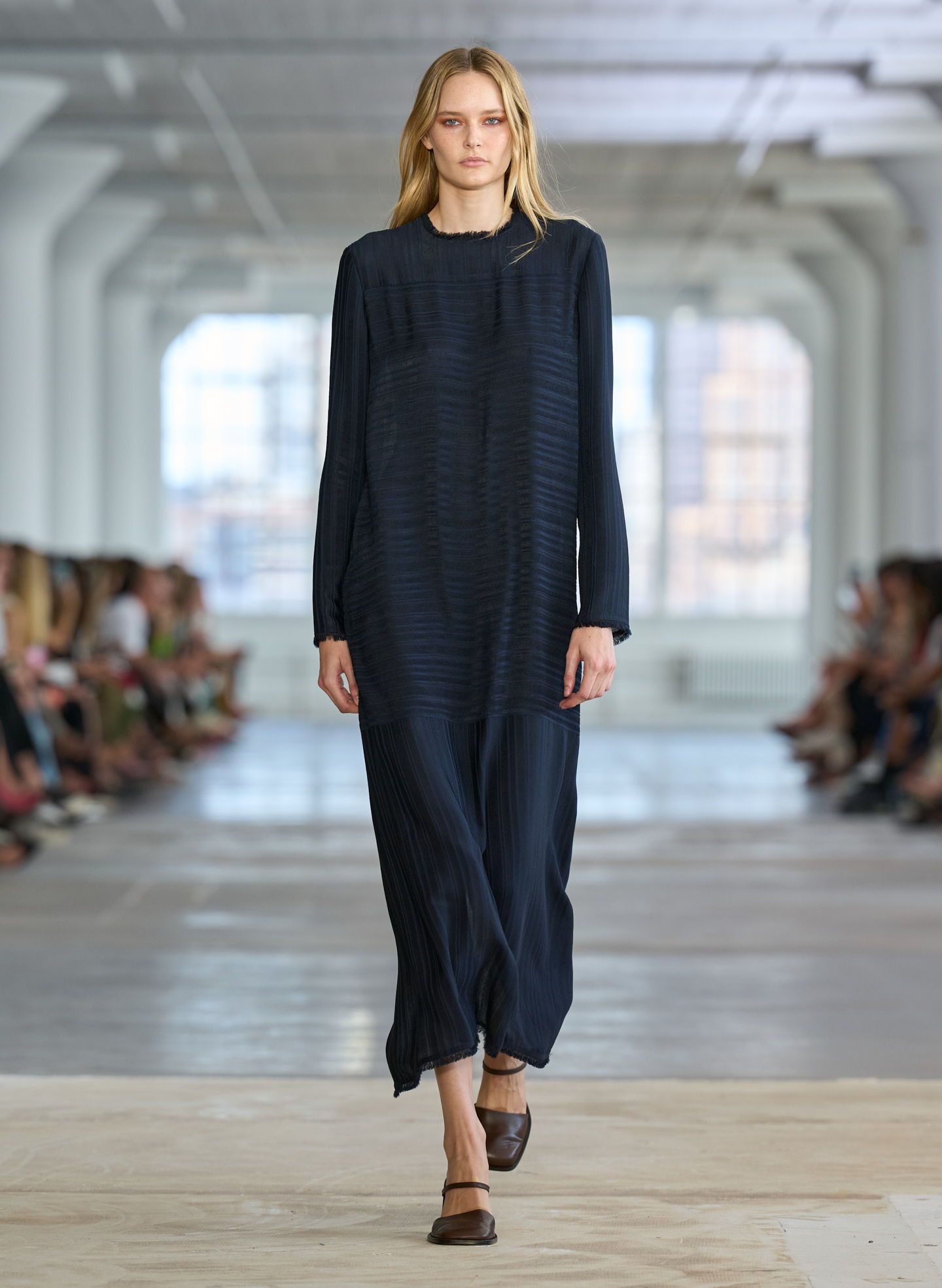 Model walking on runway wearing a long dark dress and brown shoes.